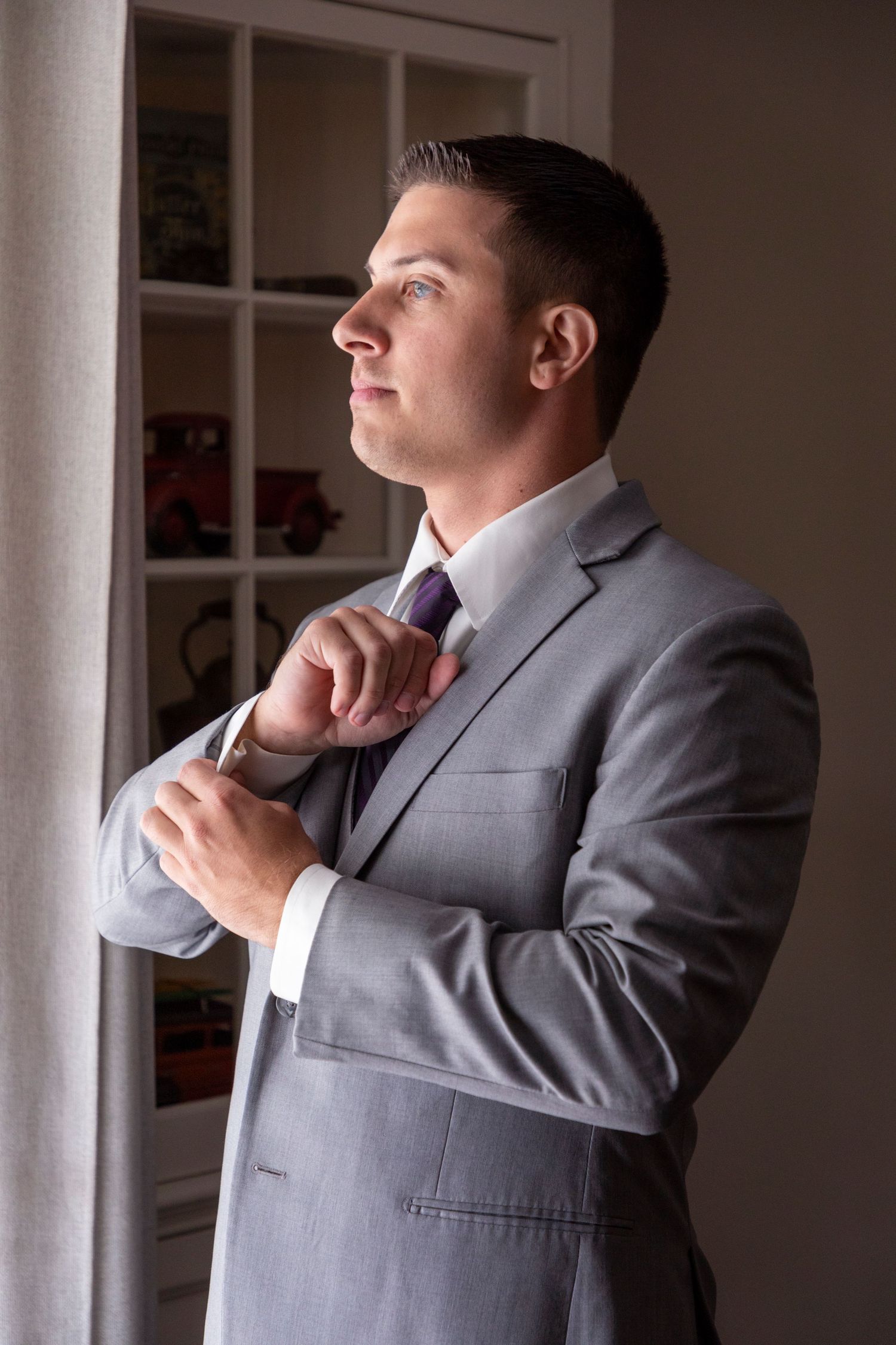 Groom in grey suit adjusts tie while getting ready in sunlit room.