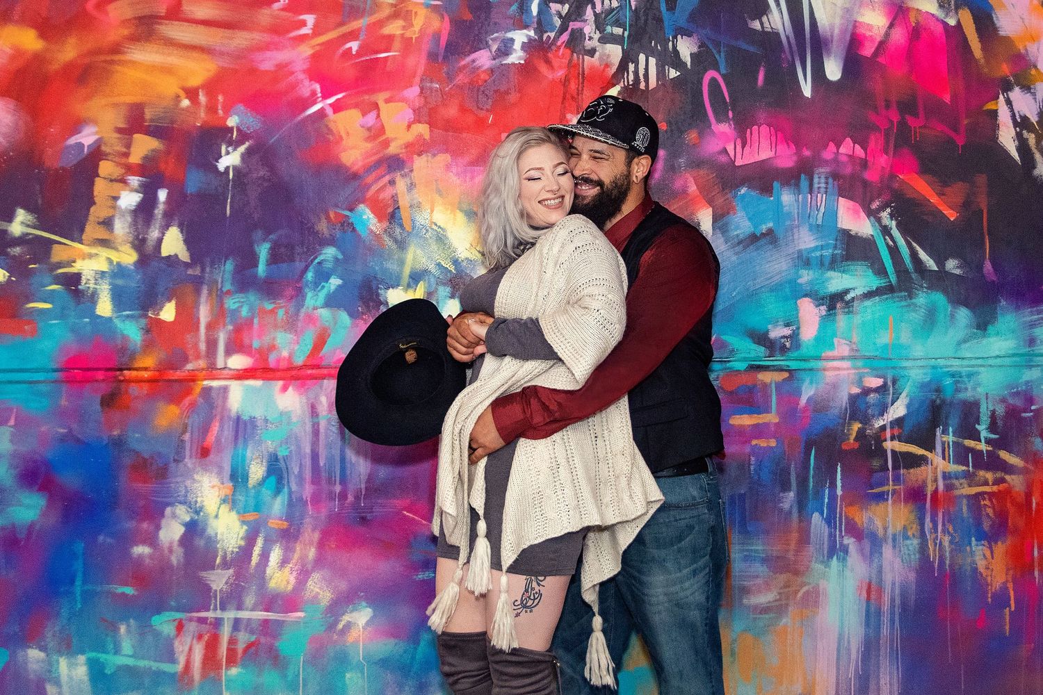 A couple embraces in front of a vibrant multicolored graffiti wall.