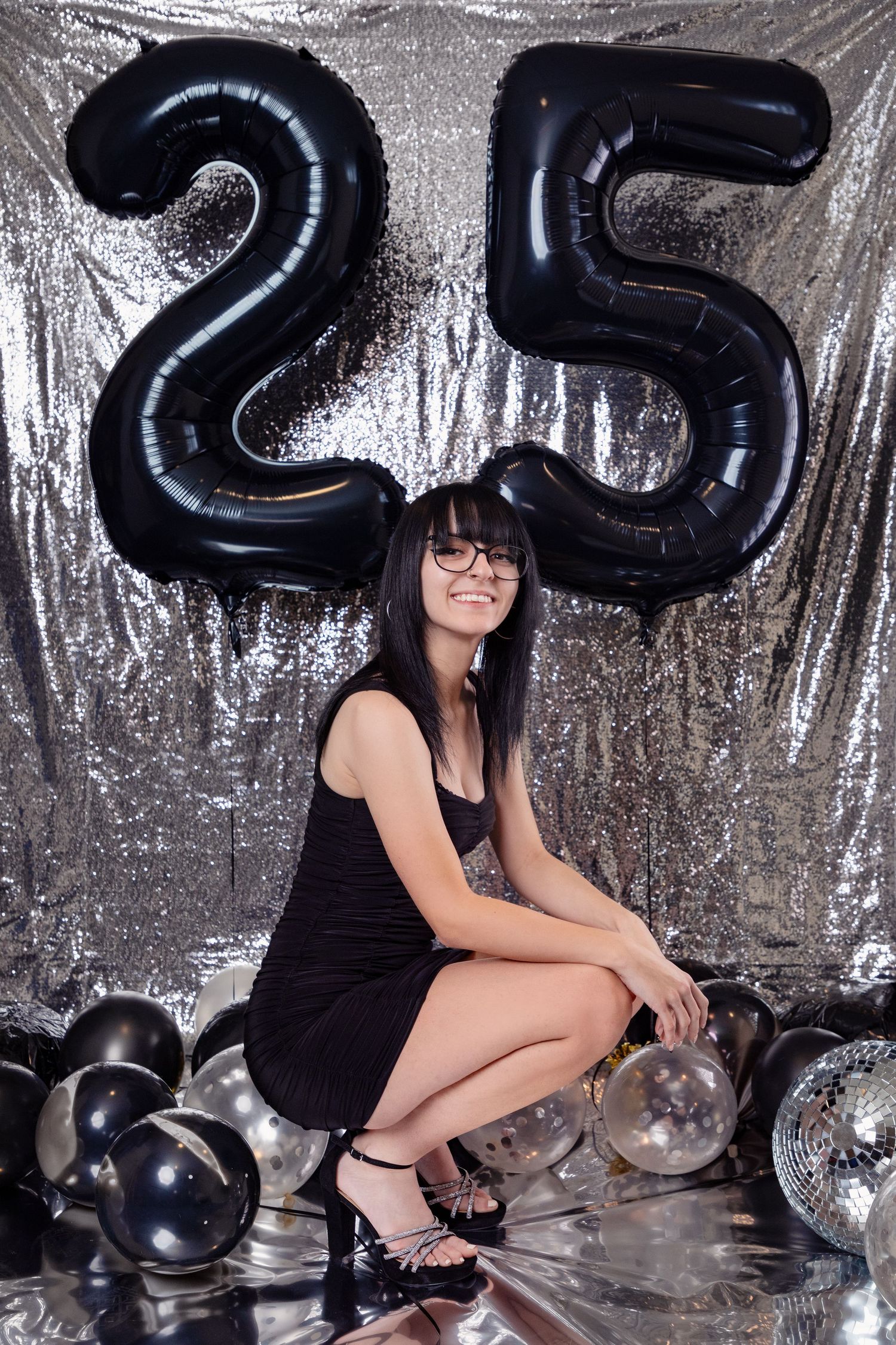 Person in black dress poses with large black number balloons against silver backdrop.