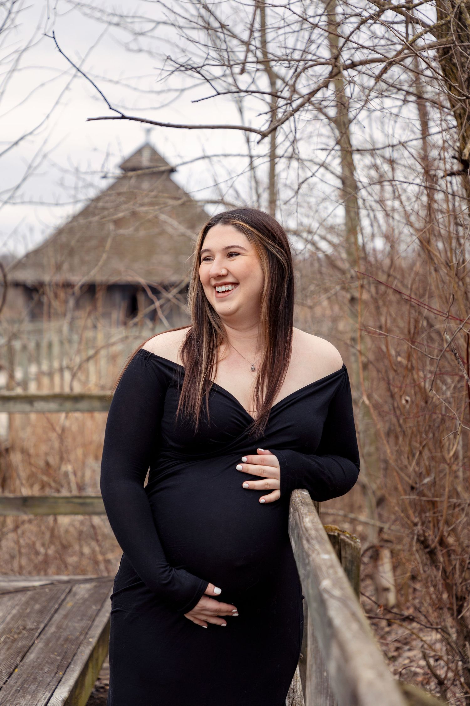 An expectant mother in a black dress laughs while posing on a wooden deck with bare winter trees.