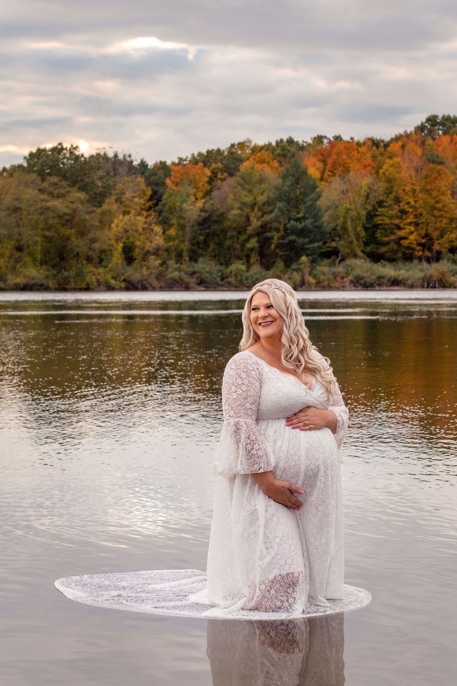 Beautiful fall maternity photos by a lake with autumn foliage in the background.