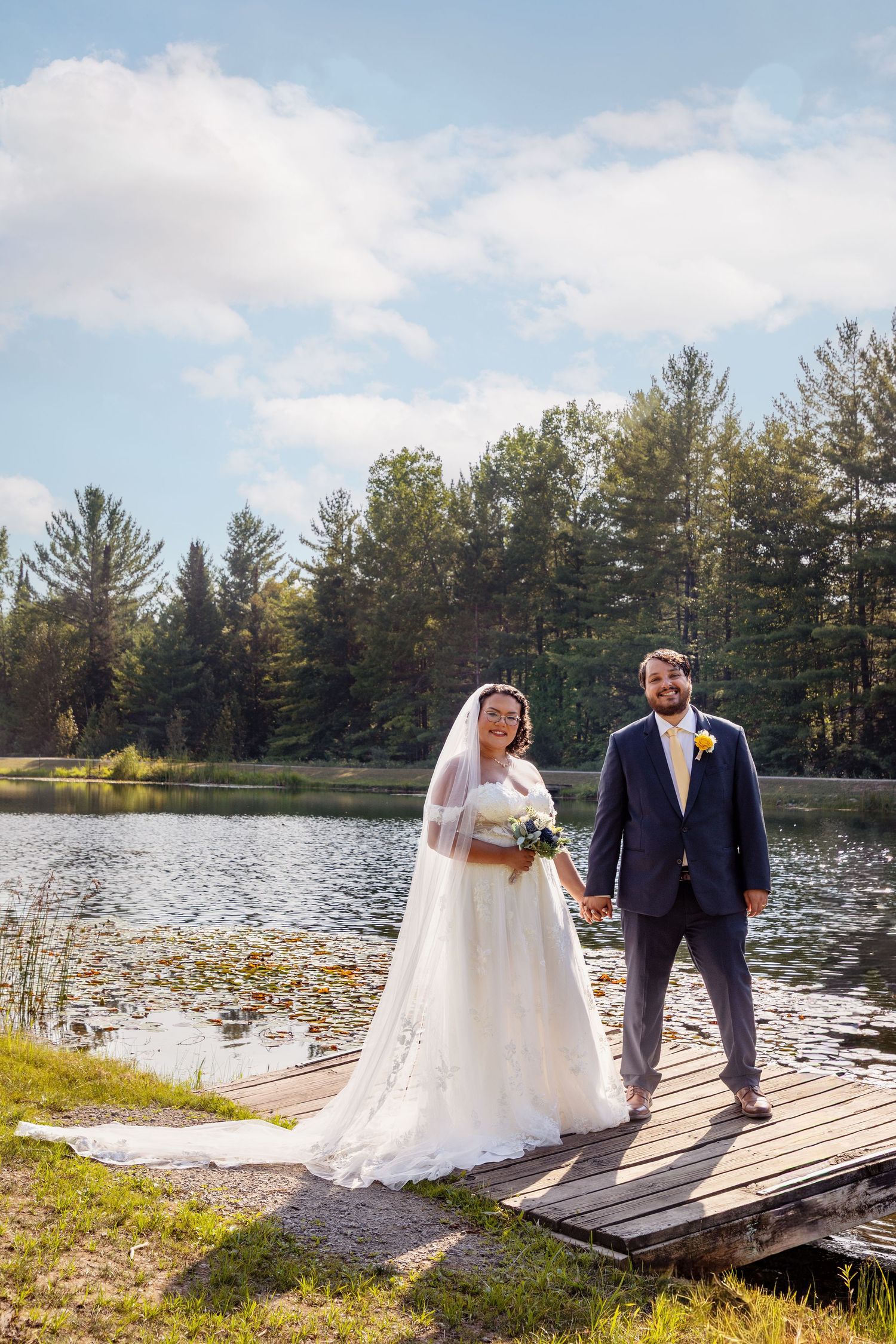 A wedding couple poses on a dock by a serene lake surrounded by pine trees.