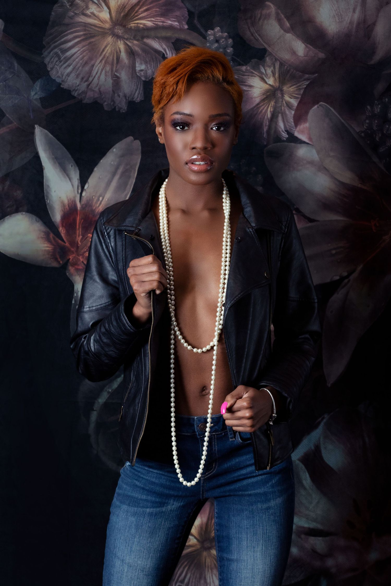 Fashion model wearing leather jacket and pearls poses against dark floral backdrop.