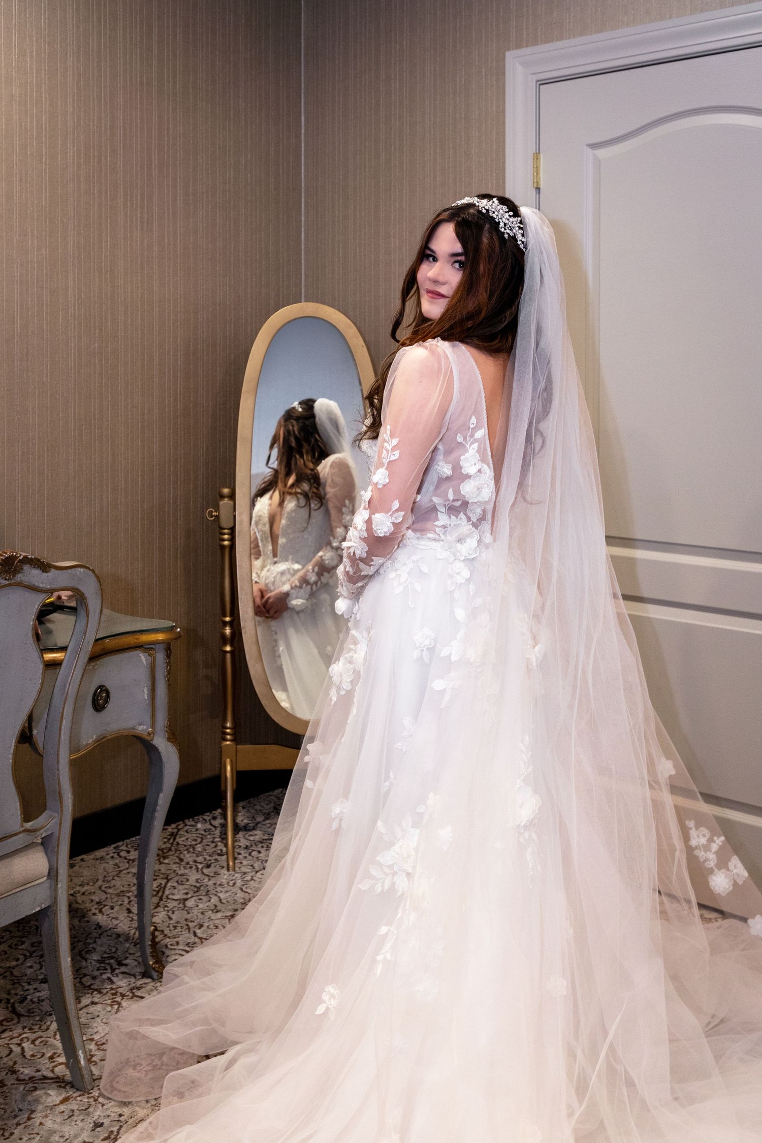 A bride in a lace wedding gown looks over her shoulder at her reflection in an ornate mirror.