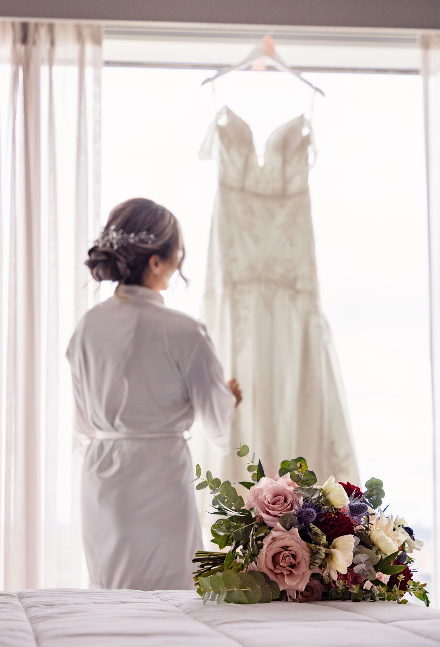 Wedding dress hangs in window with delicate pink and purple floral bouquet arrangement on table.