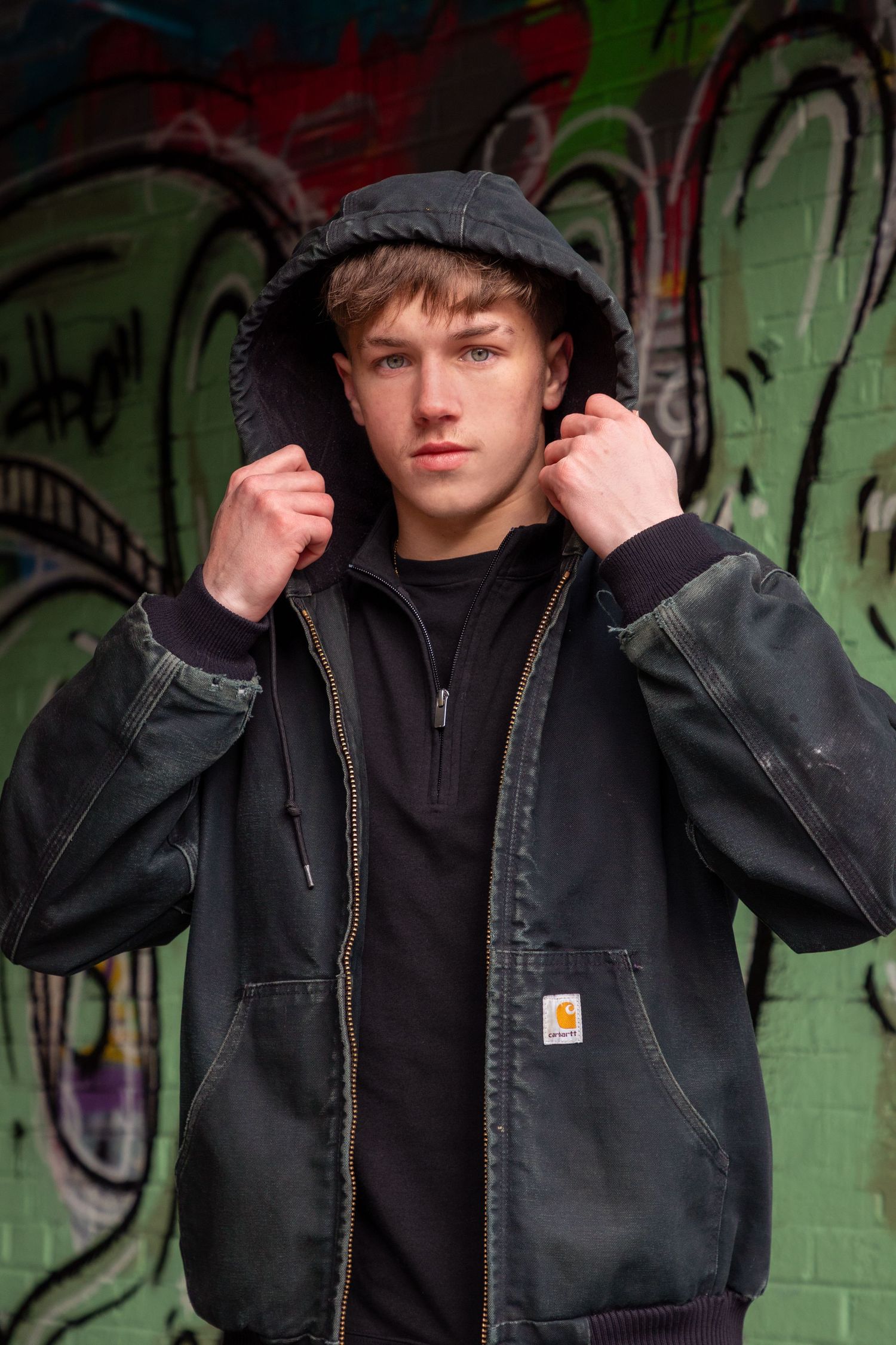 Person in black Carhartt jacket poses against graffiti wall.