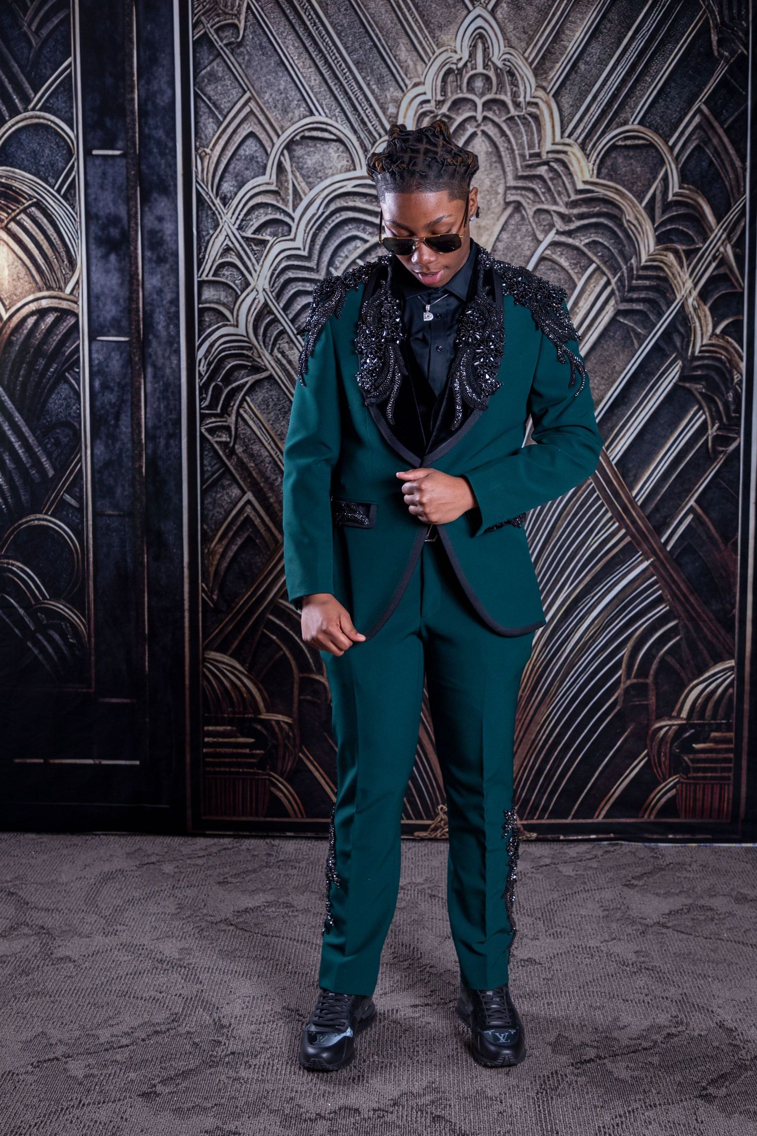 Stylish poses in an emerald green suit against a decorative dark background.