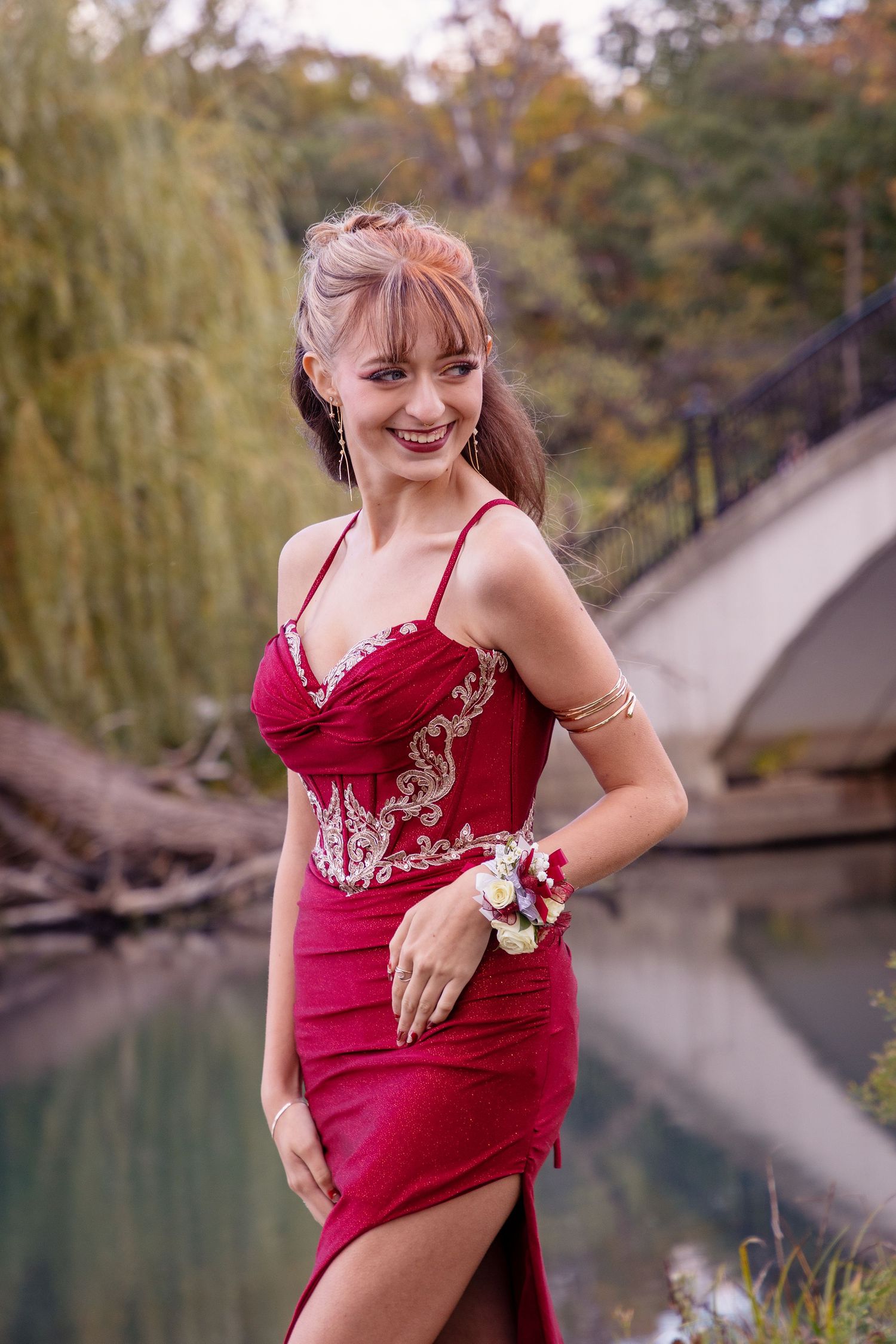 Person in red dress poses playfully by lake with autumn foliage in background.