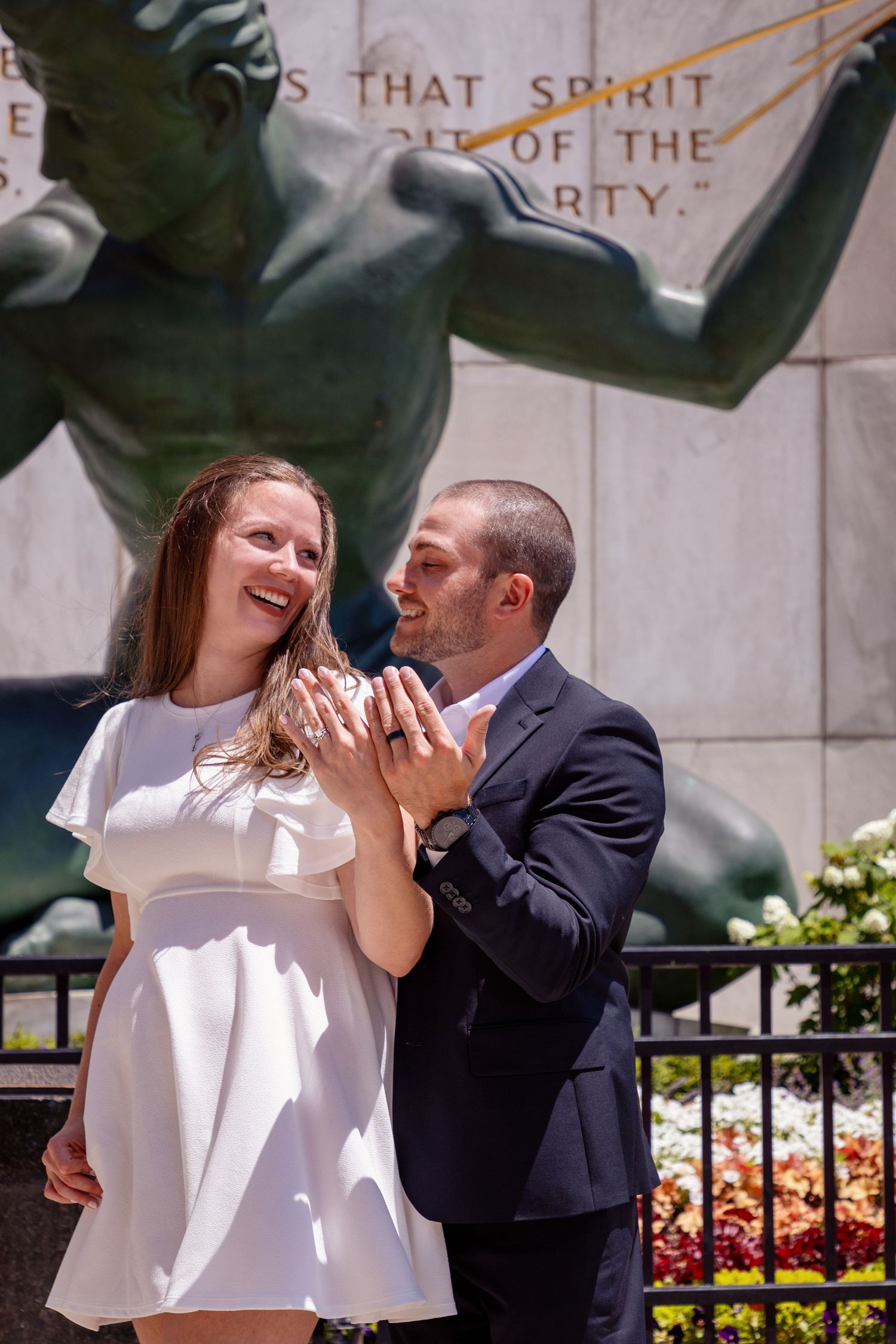 A couple embraces near a decorative railing with flowers and stone architecture.