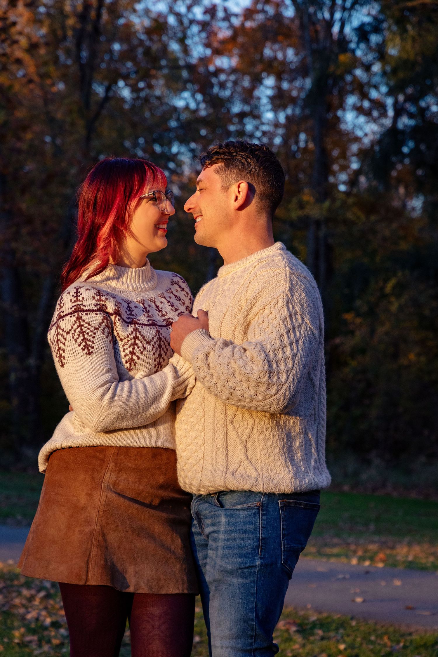 Couple in cozy sweaters embracing during sunset with autumn foliage in the background.