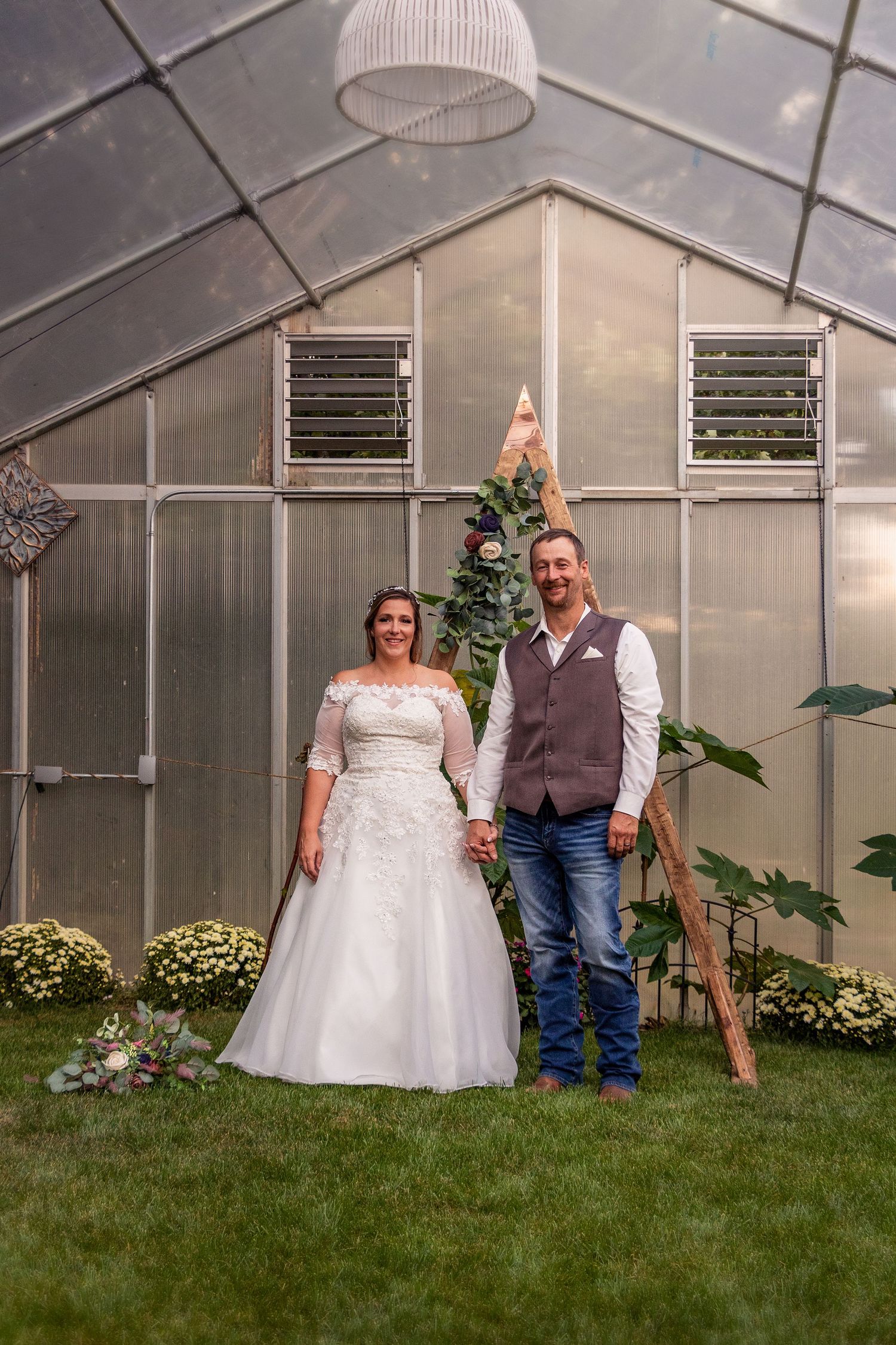 A couple poses for a wedding photo inside a greenhouse with decorative lighting.