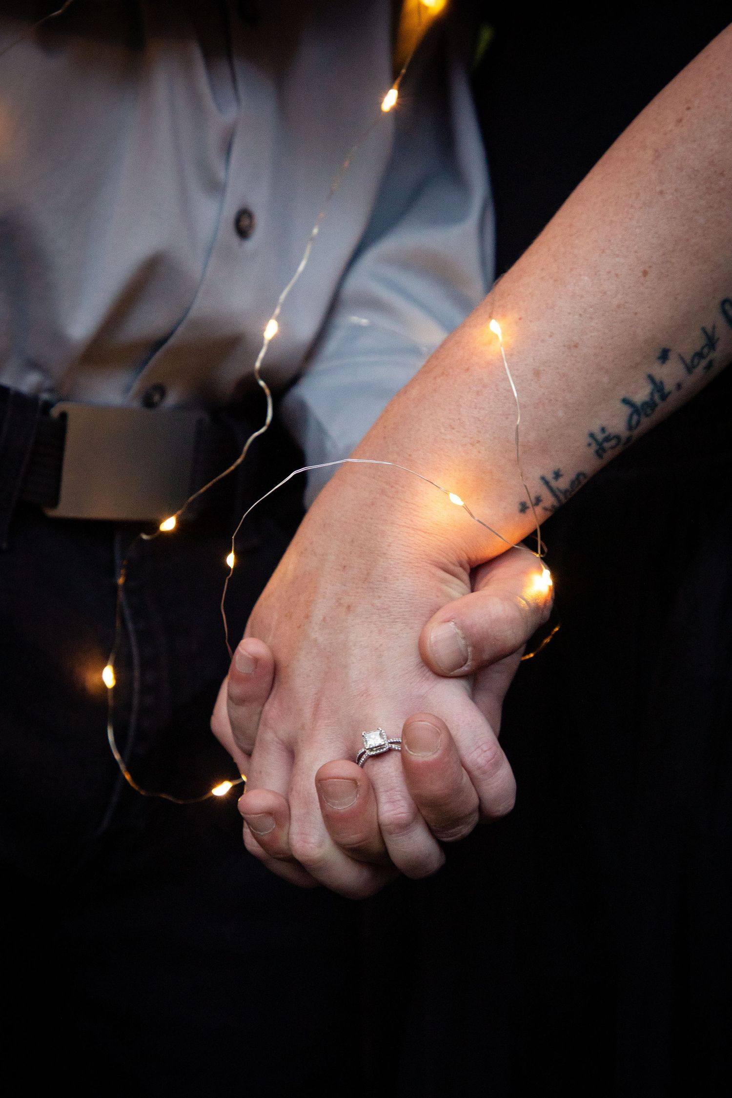 Close-up of clasped hands wrapped in twinkling fairy lights showing an engagement ring.