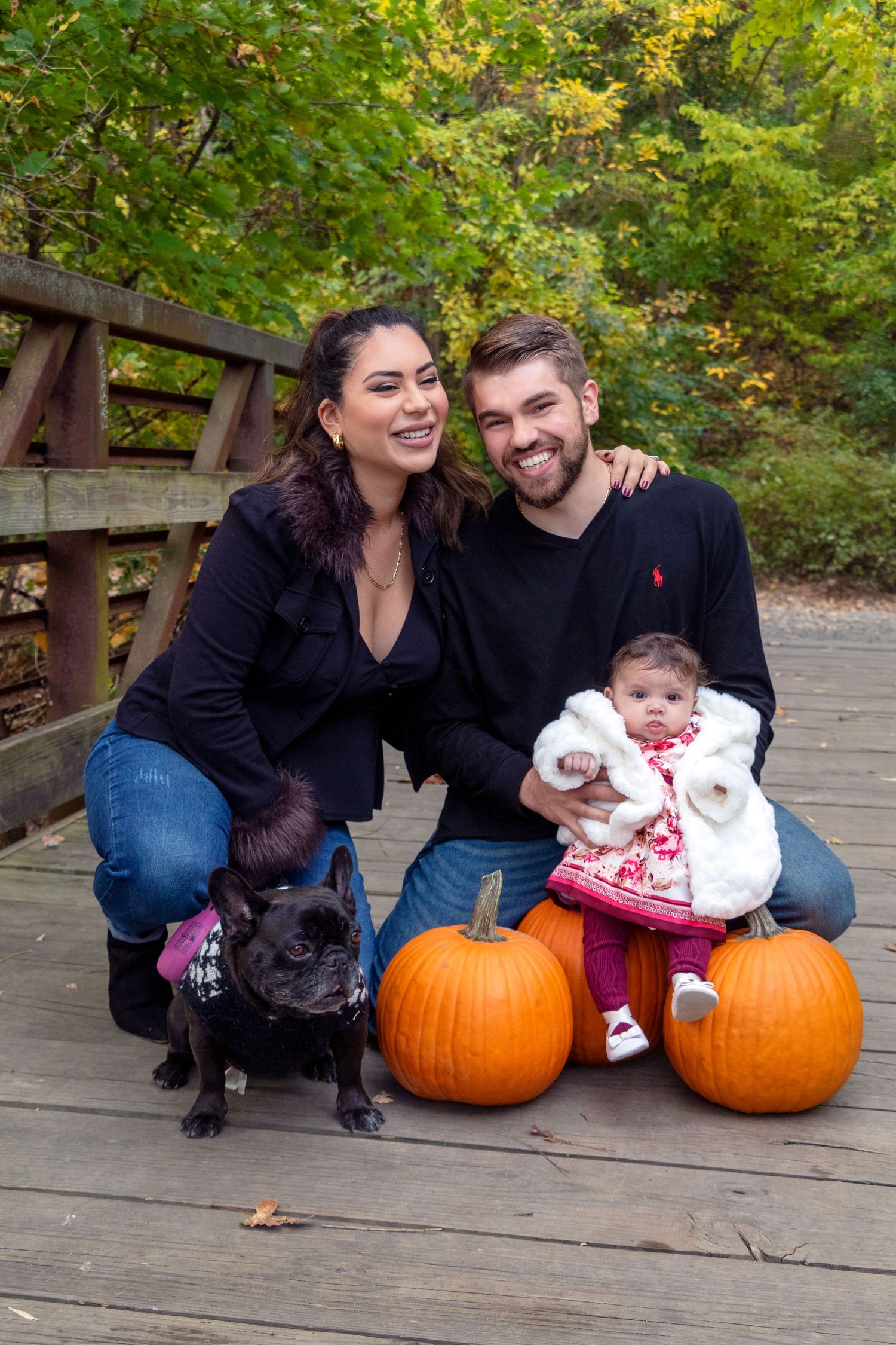 Family poses with pumpkins and a dog on a wooden deck during fall season.