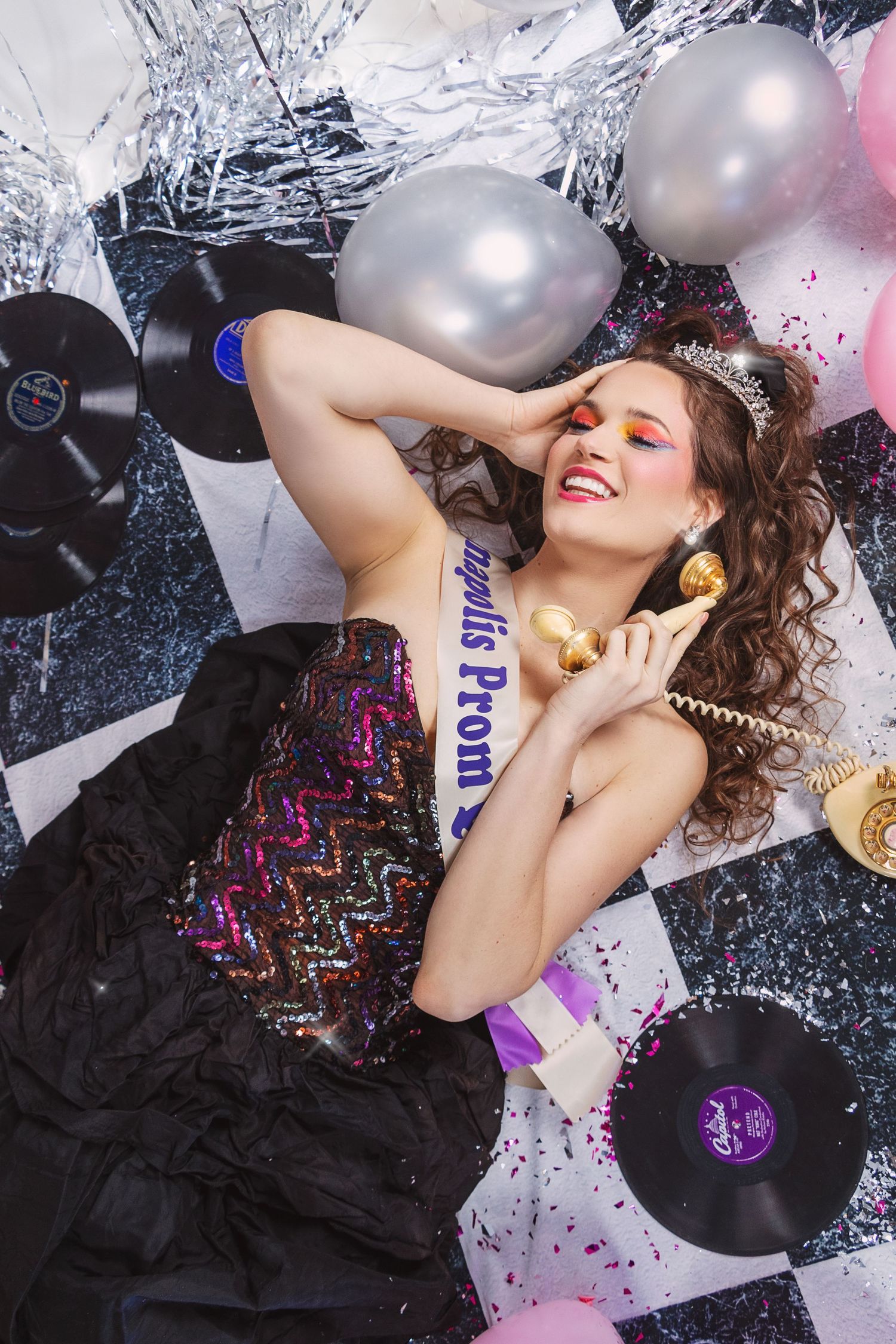 Fashion model lying on floor with silver balloons and confetti in glamorous party setting.