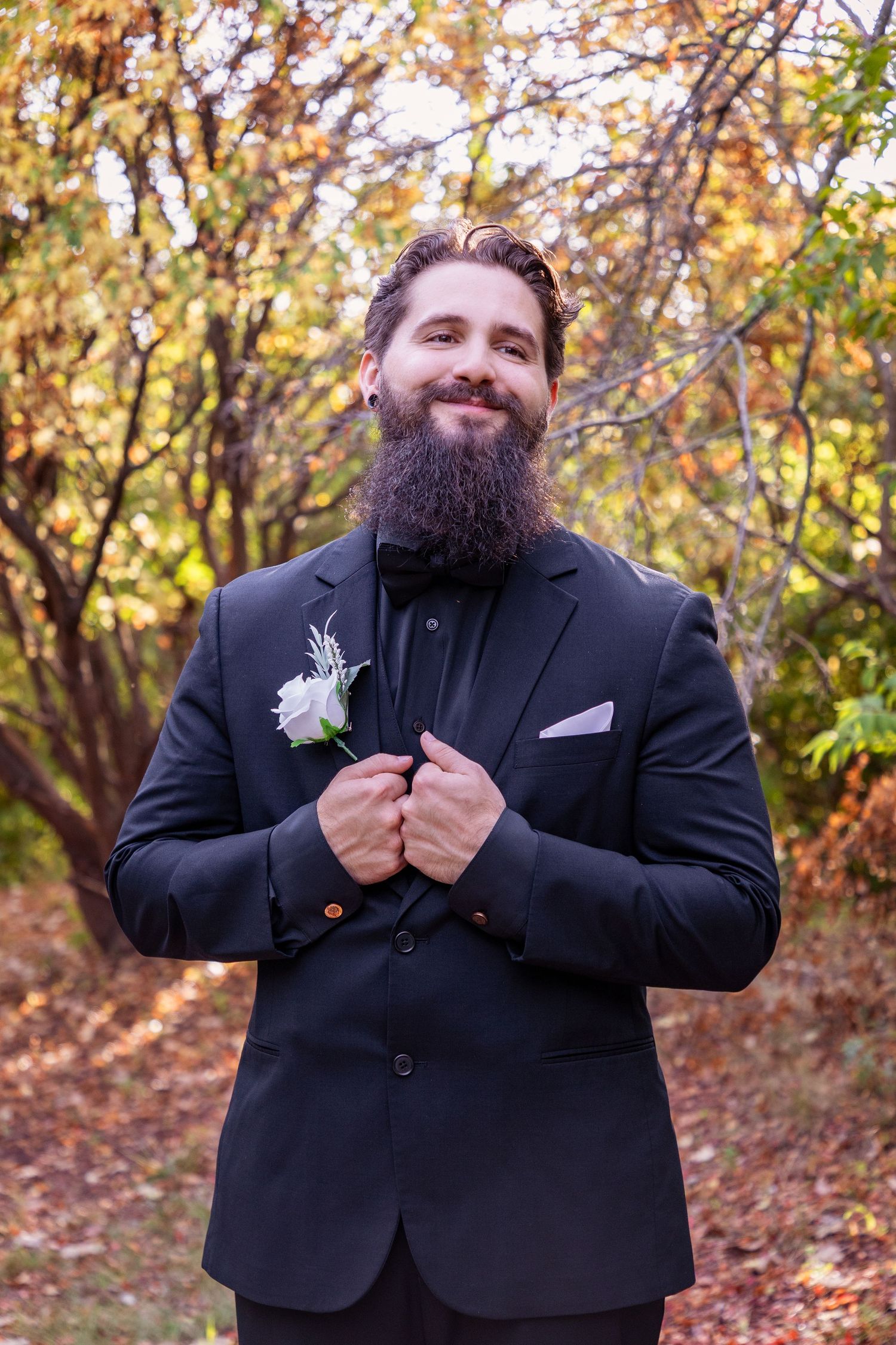 Groom in black suit with boutonniere smiles in autumn foliage.