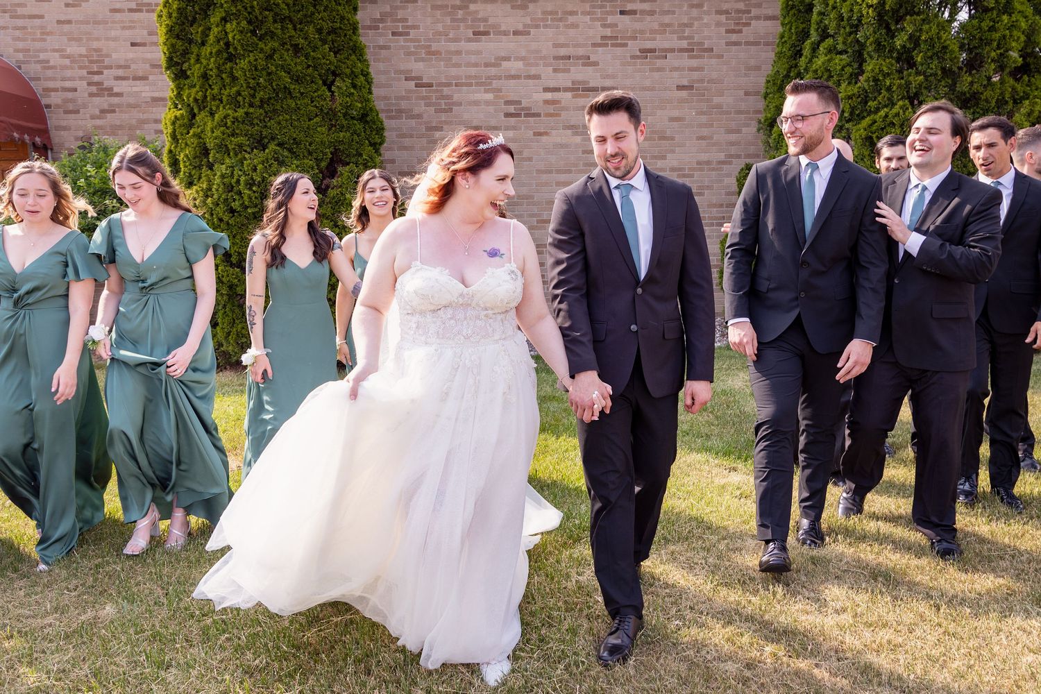 Large wedding party walks together on grounds wearing sage green and navy attire.
