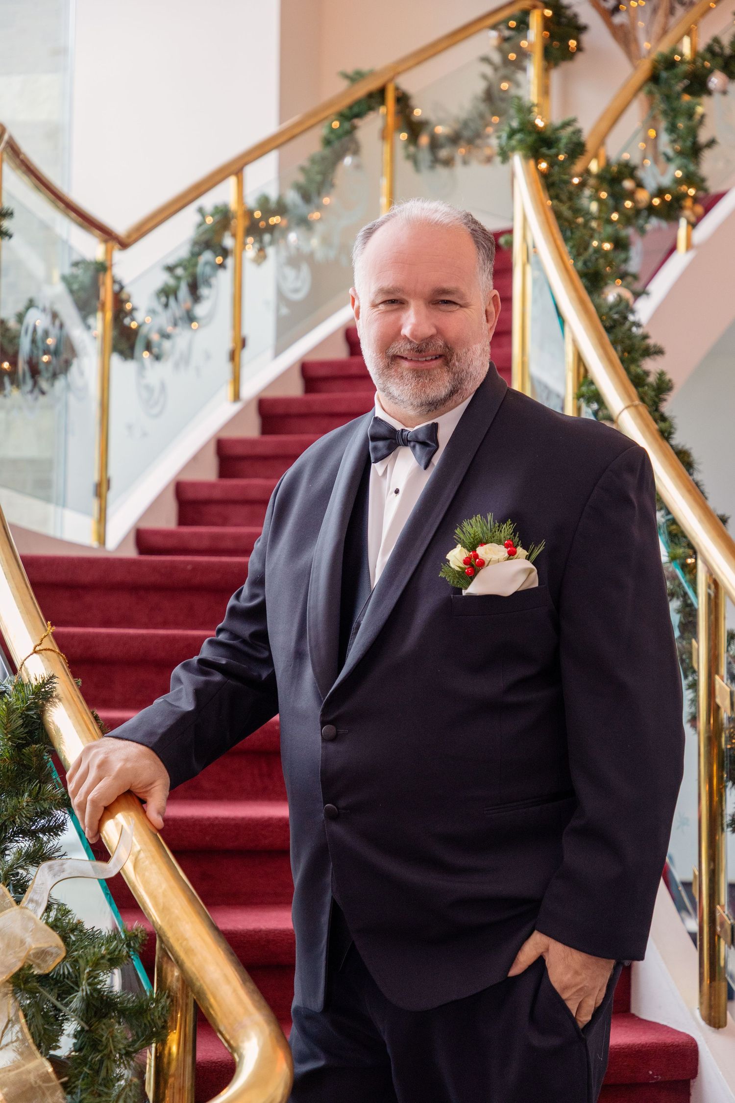 Formal portraits of a groom in a black tuxedo posing on an elegant staircase decorated with holiday garland.
