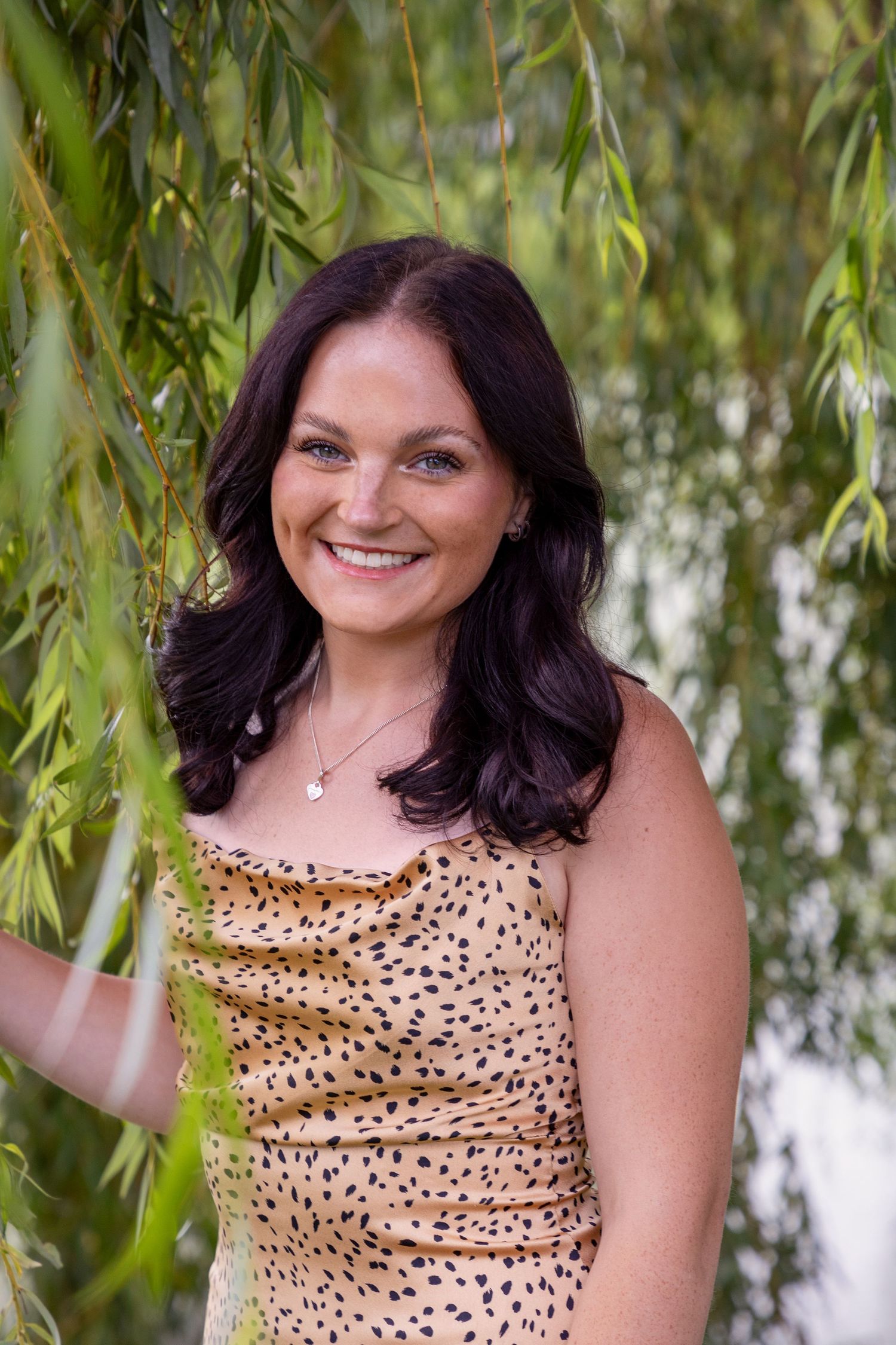 Woman in leopard print top poses under willow tree branches near water.