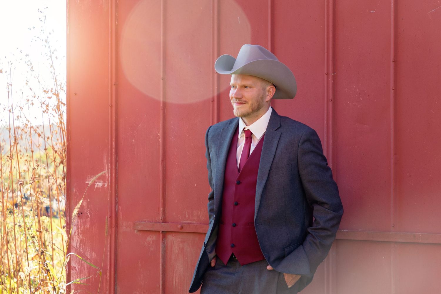 Stylish man in western attire leans against red barn wall wearing cowboy hat.