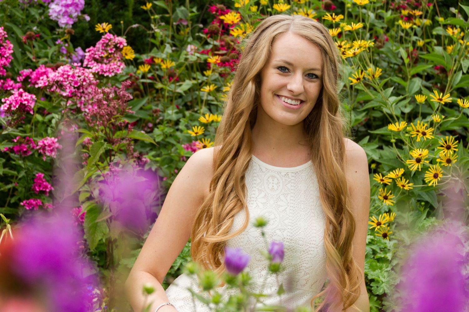 Woman in white dress poses in garden with colorful pink and yellow flowers.