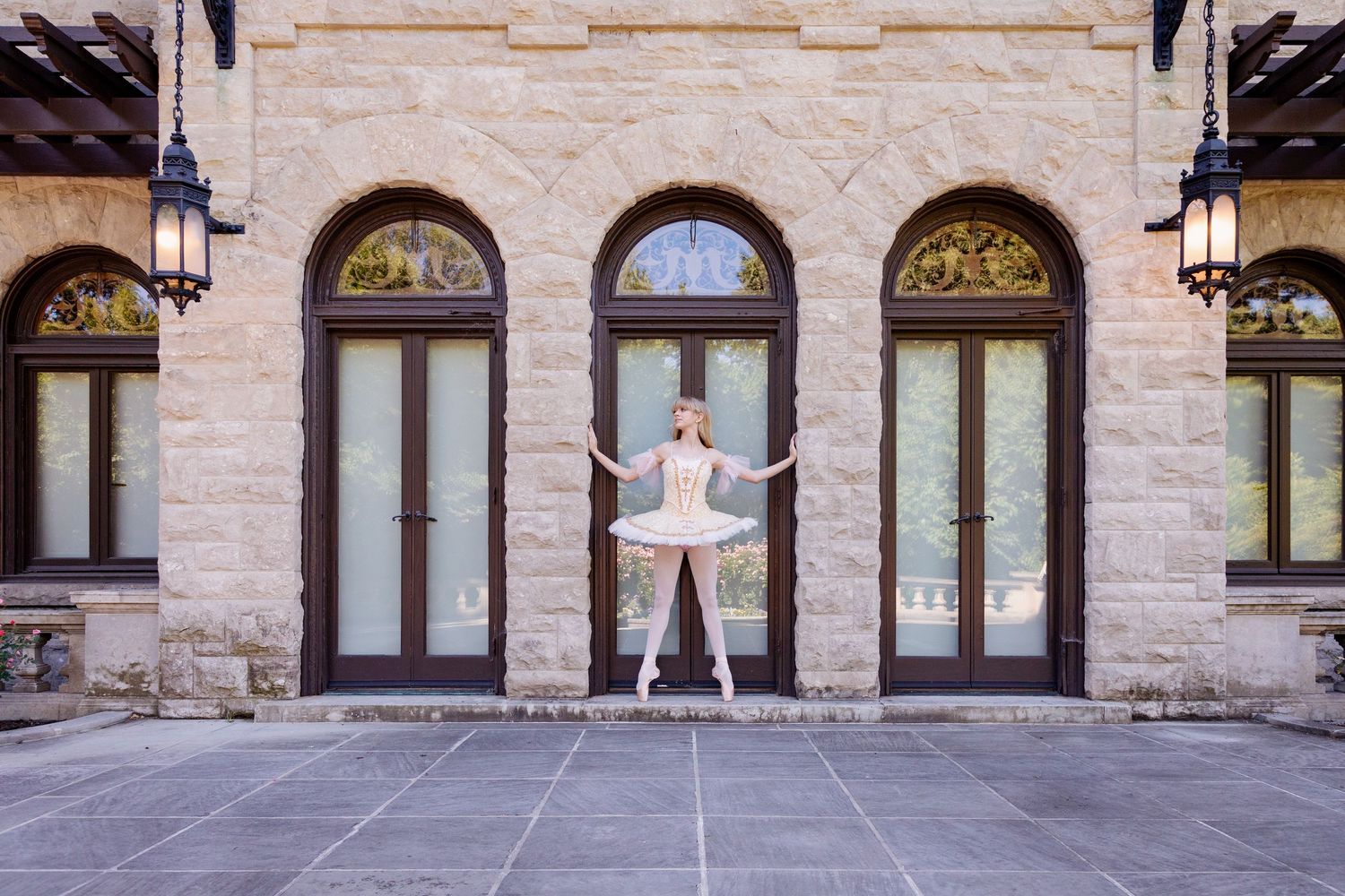 A ballet dancer in white tutu poses between arched doorways of a stone building.