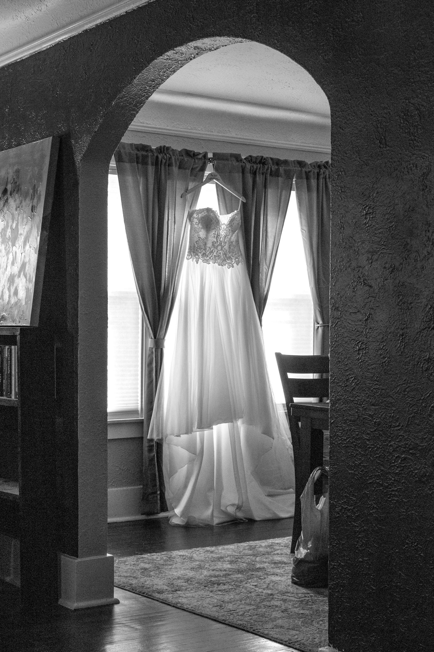 An elegant white wedding dress hangs in an arched doorway near curtained windows.