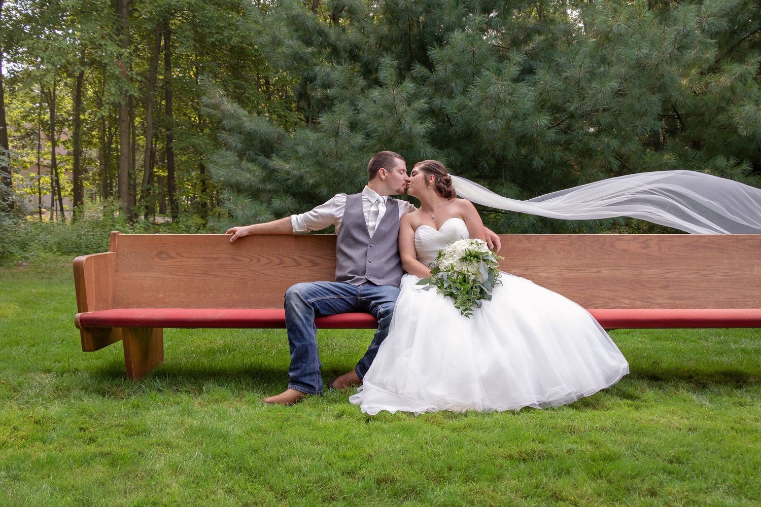 Couple in wedding attire reclining on a wooden bench in an outdoor garden setting.