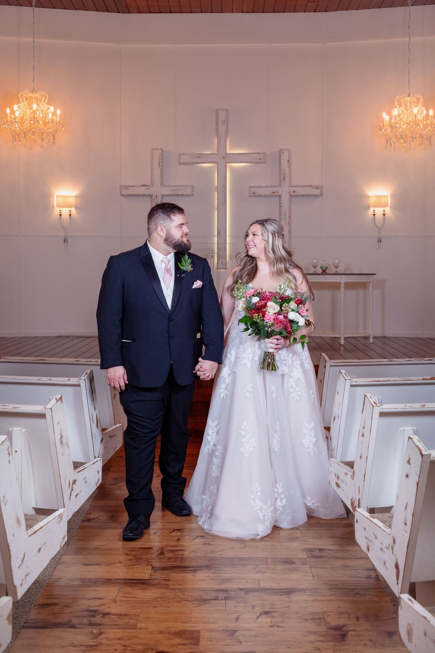 Wedding couple walks through white church sanctuary with wooden pews and crystal chandeliers.