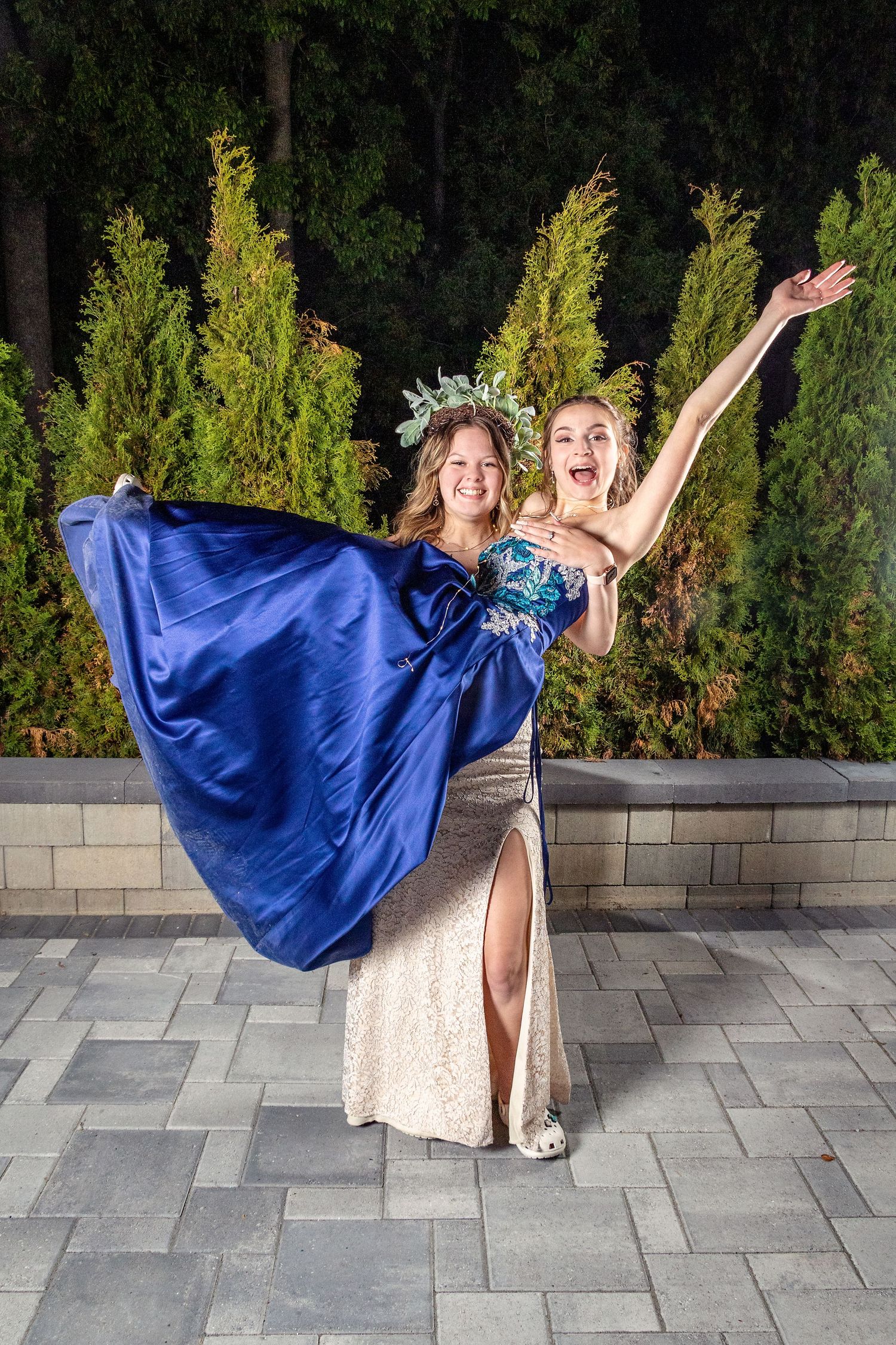 Two people in formal attire strike a dramatic pose against an evening backdrop of evergreen trees.