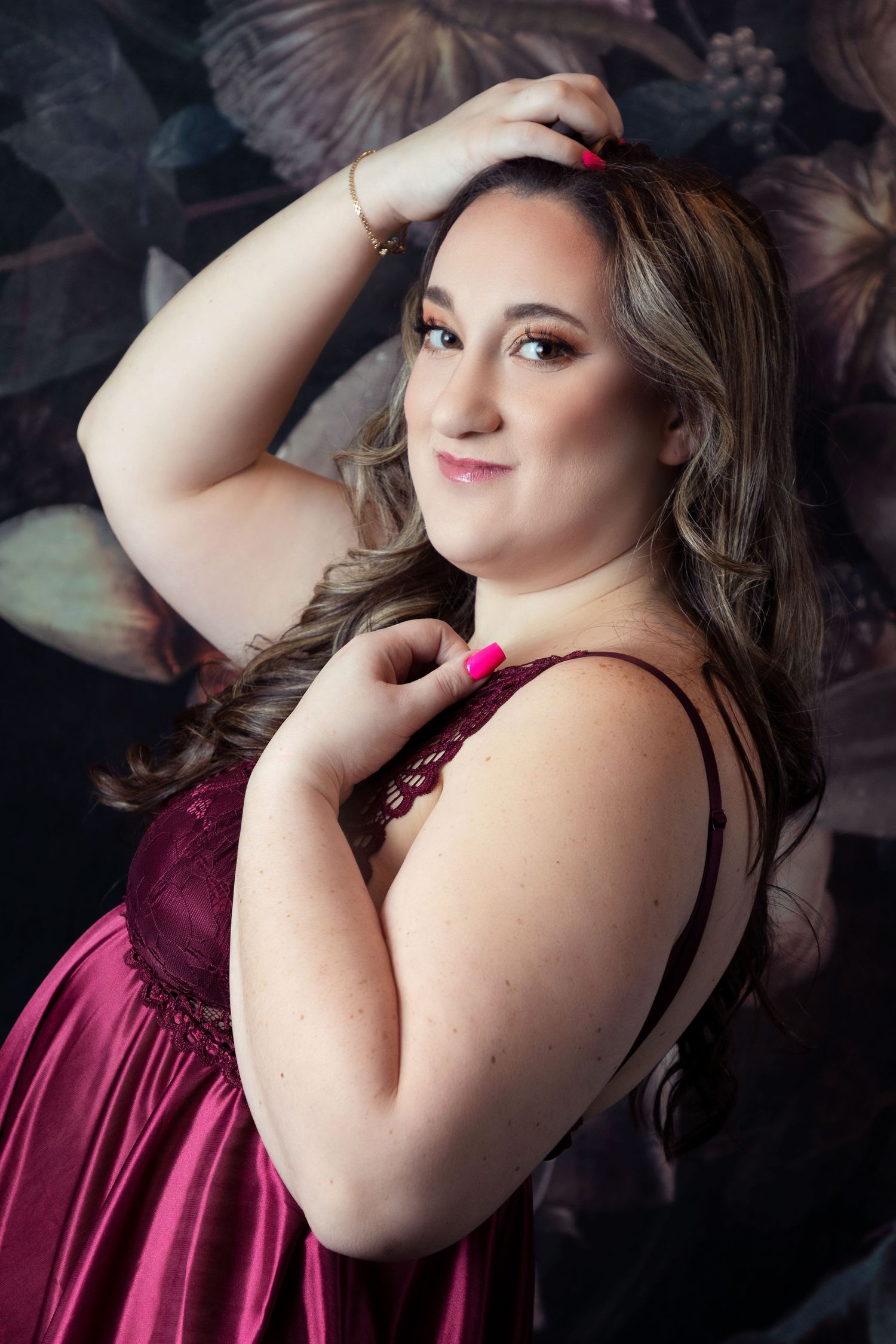 Portrait of someone in a burgundy dress posing against a dark floral backdrop.