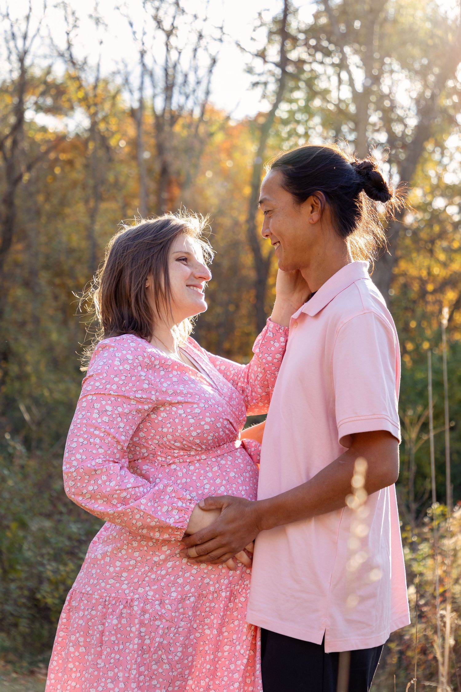 A romantic maternity photoshoot featuring a couple in pink outfits against an autumn forest backdrop.