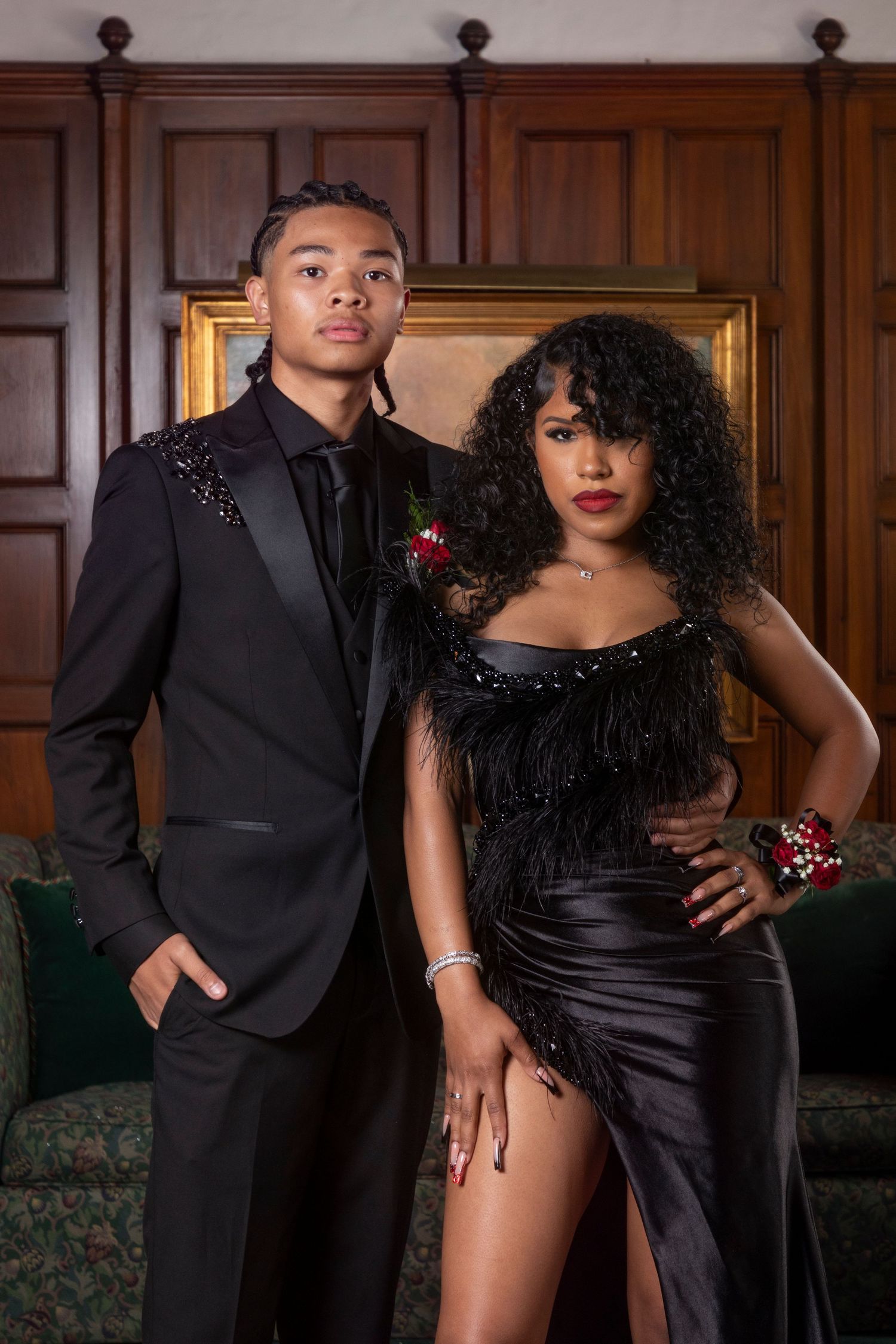 Elegant couple in formal black attire pose together against wood-paneled wall at an event.