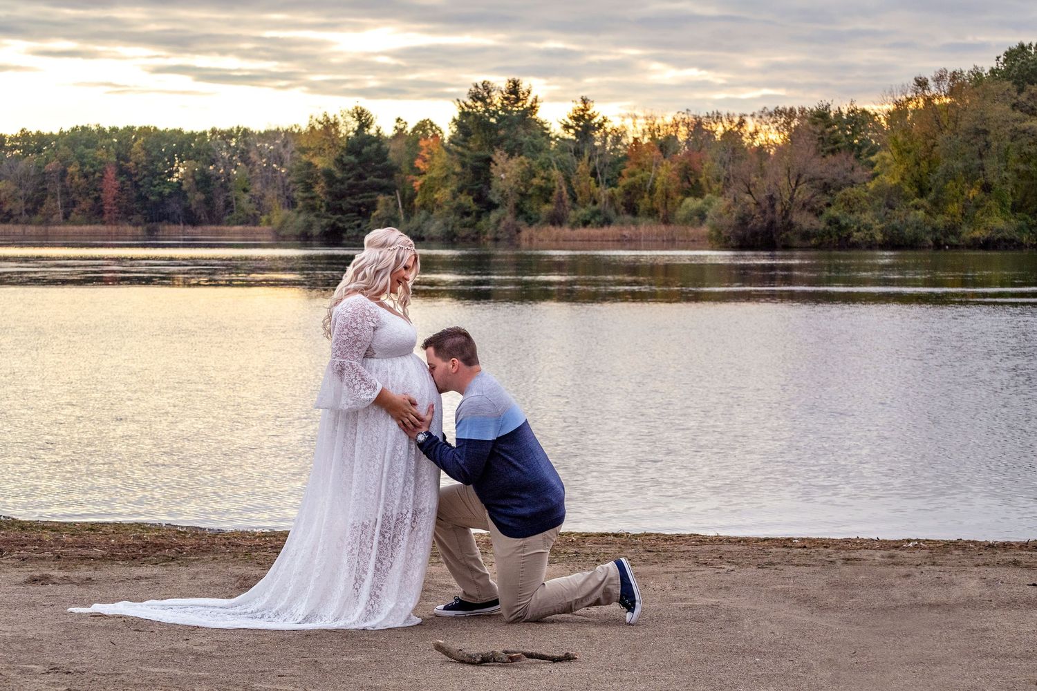 Sunset maternity photos by a serene lake with fall foliage in the background.