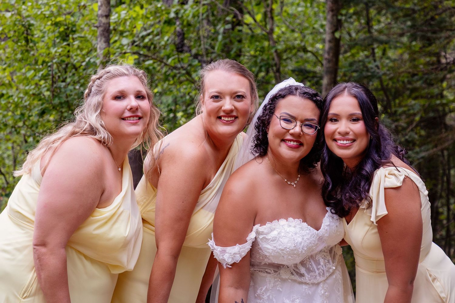 Bridesmaids in yellow dresses share a joyful moment in a forest setting.