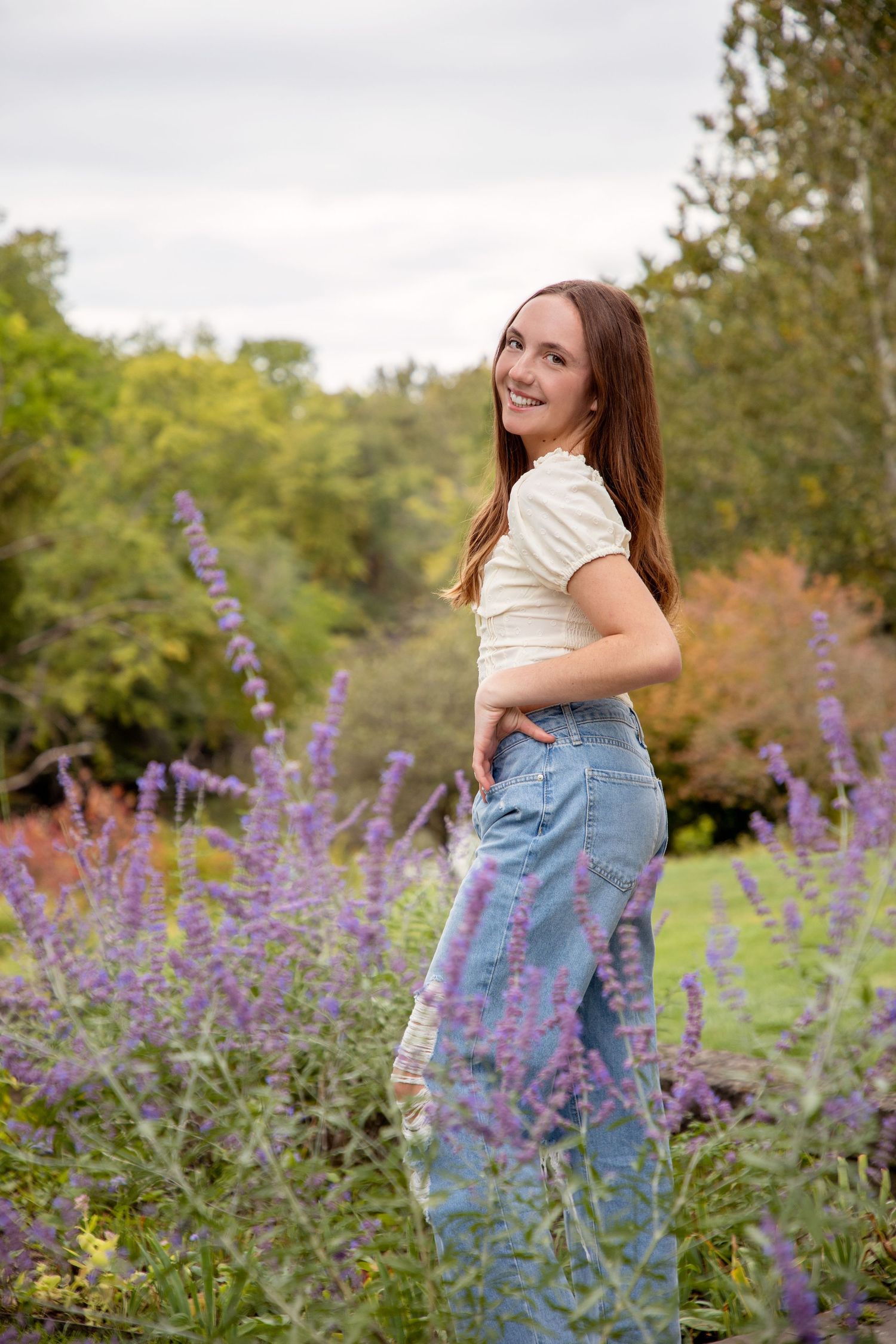 Person in casual outfit poses playfully among purple wildflowers in natural setting.