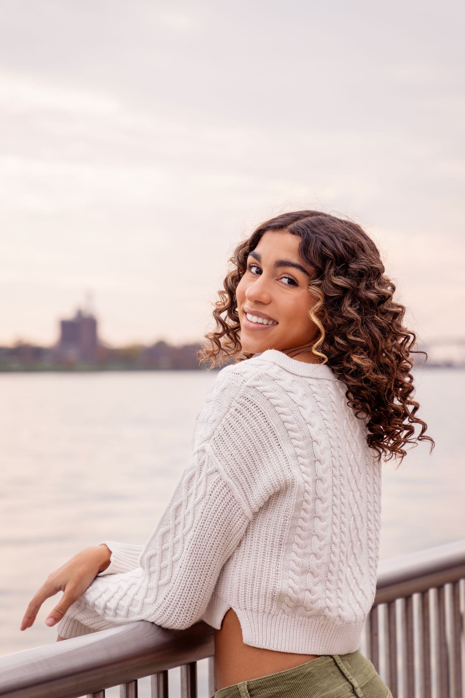 Woman in white knit sweater poses by waterfront railing at sunset.