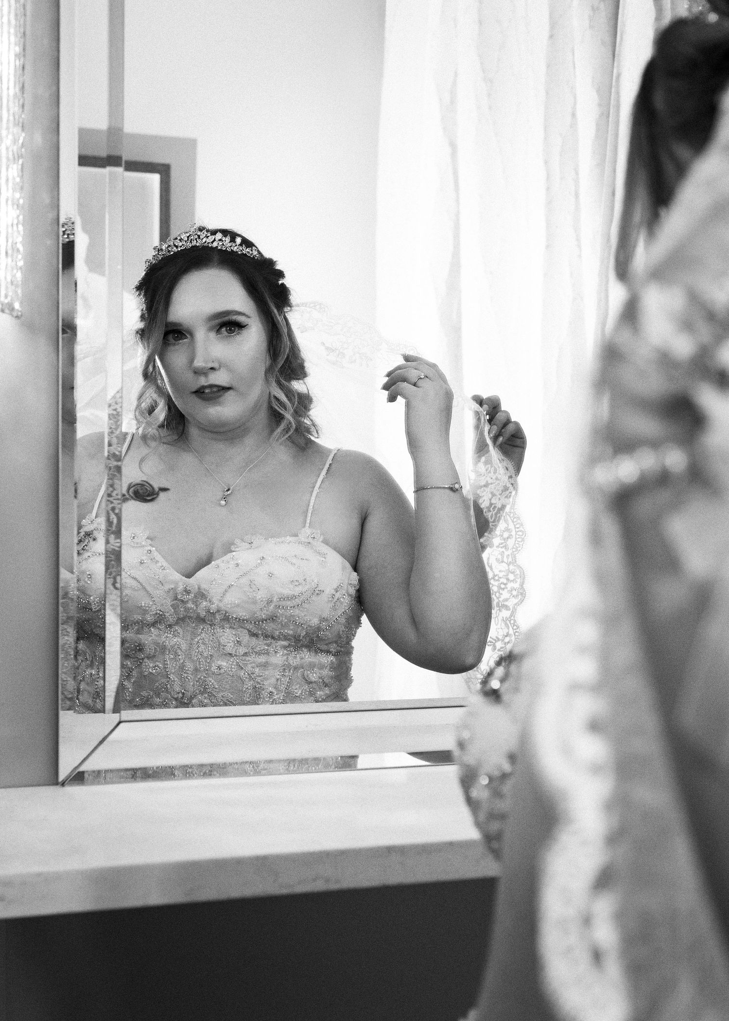 Bride in lace dress looks at reflection in elegant white-framed mirror.