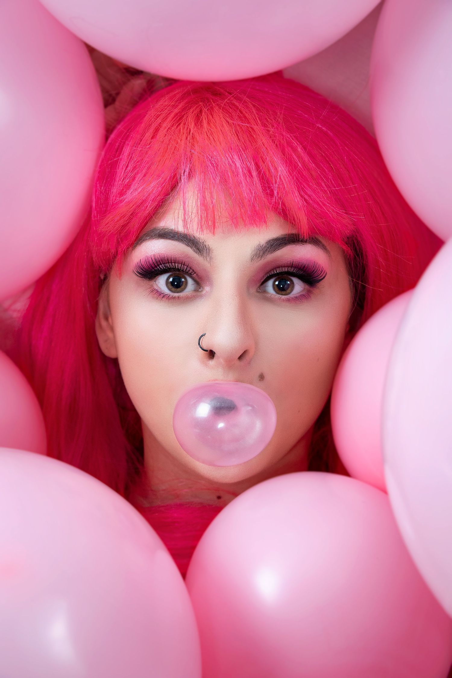 Pink-themed portrait with bright hair blowing bubble gum surrounded by pink balloons.