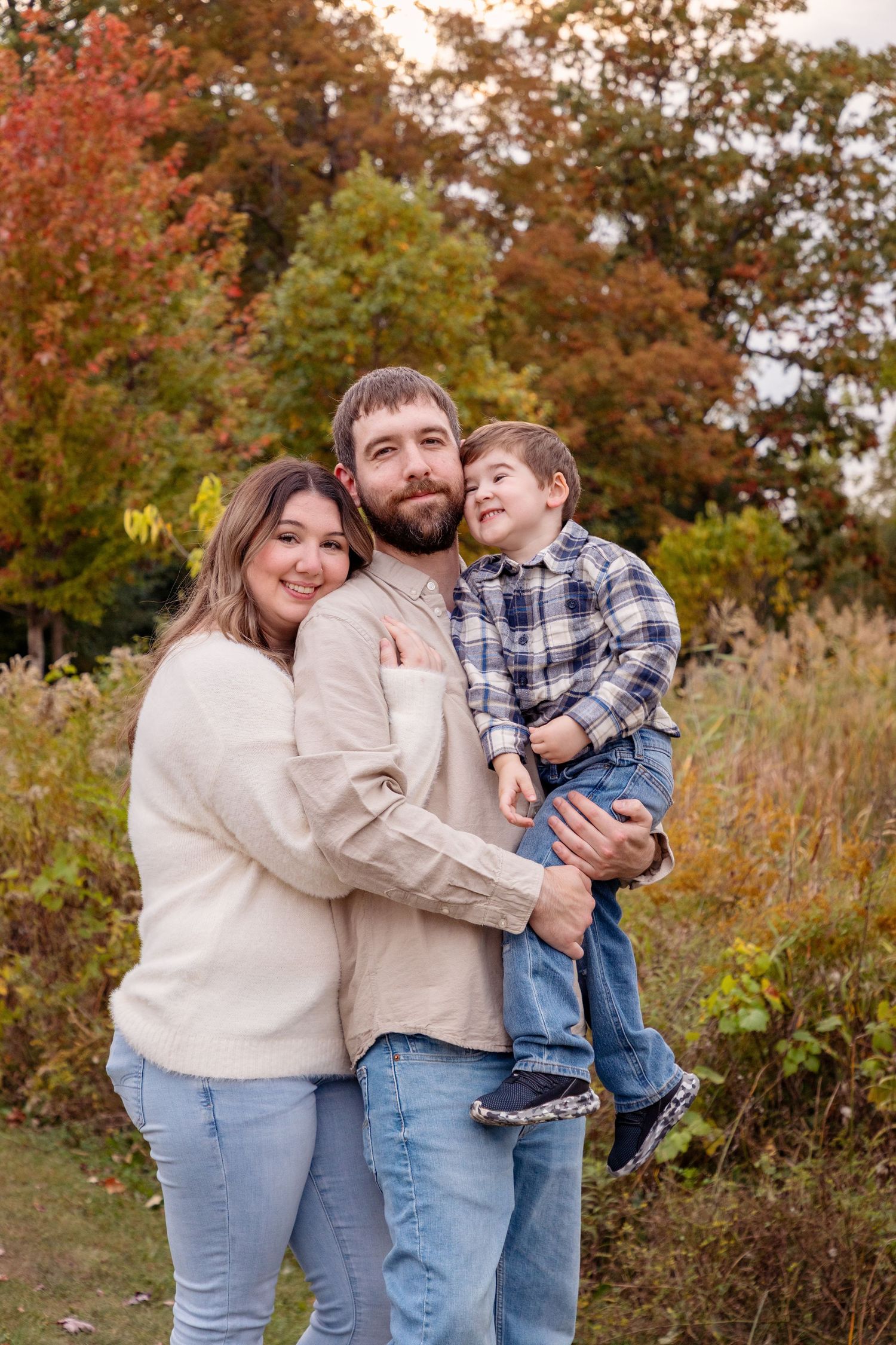 Family wearing coordinated cream sweaters and jeans pose together against autumn trees.