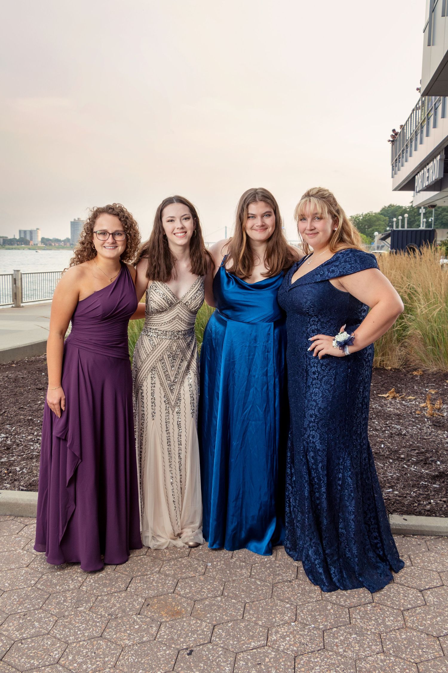 Four women in elegant formal gowns pose together on a waterfront terrace.