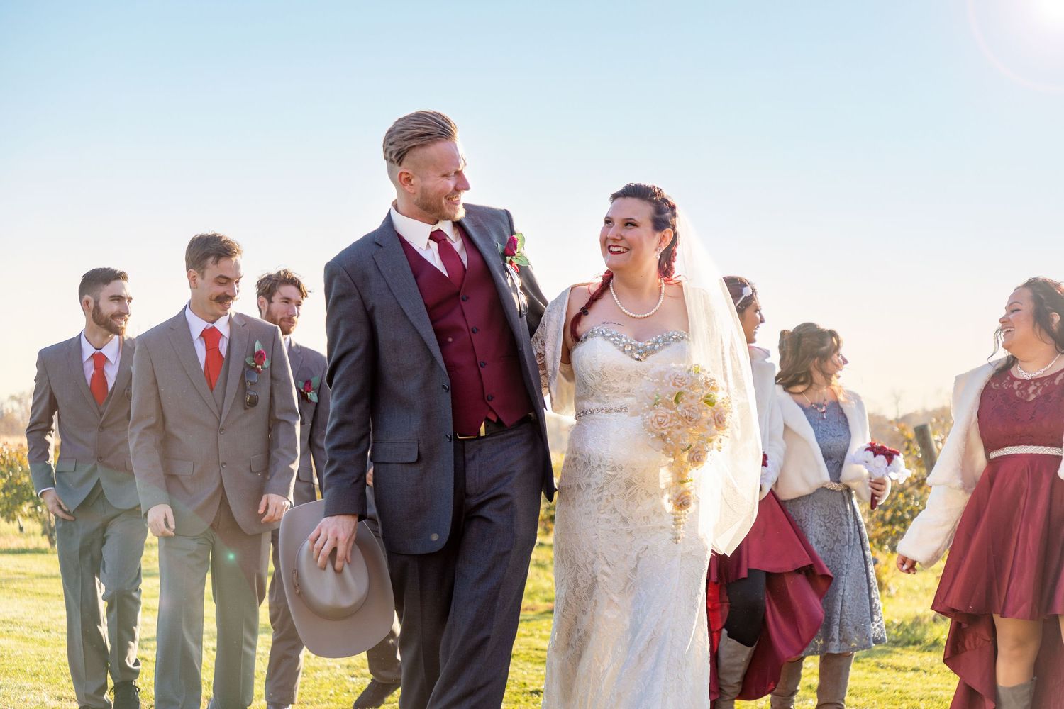 Wedding party in burgundy and gray attire poses together outdoors on a sunny day.