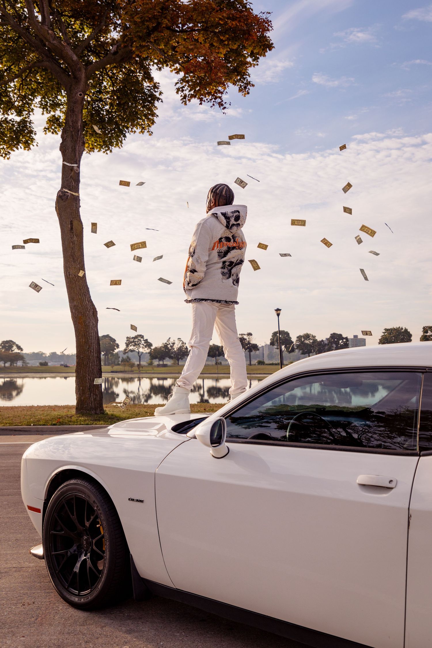 A white Dodge Challenger with someone standing on top while money falls from the sky.