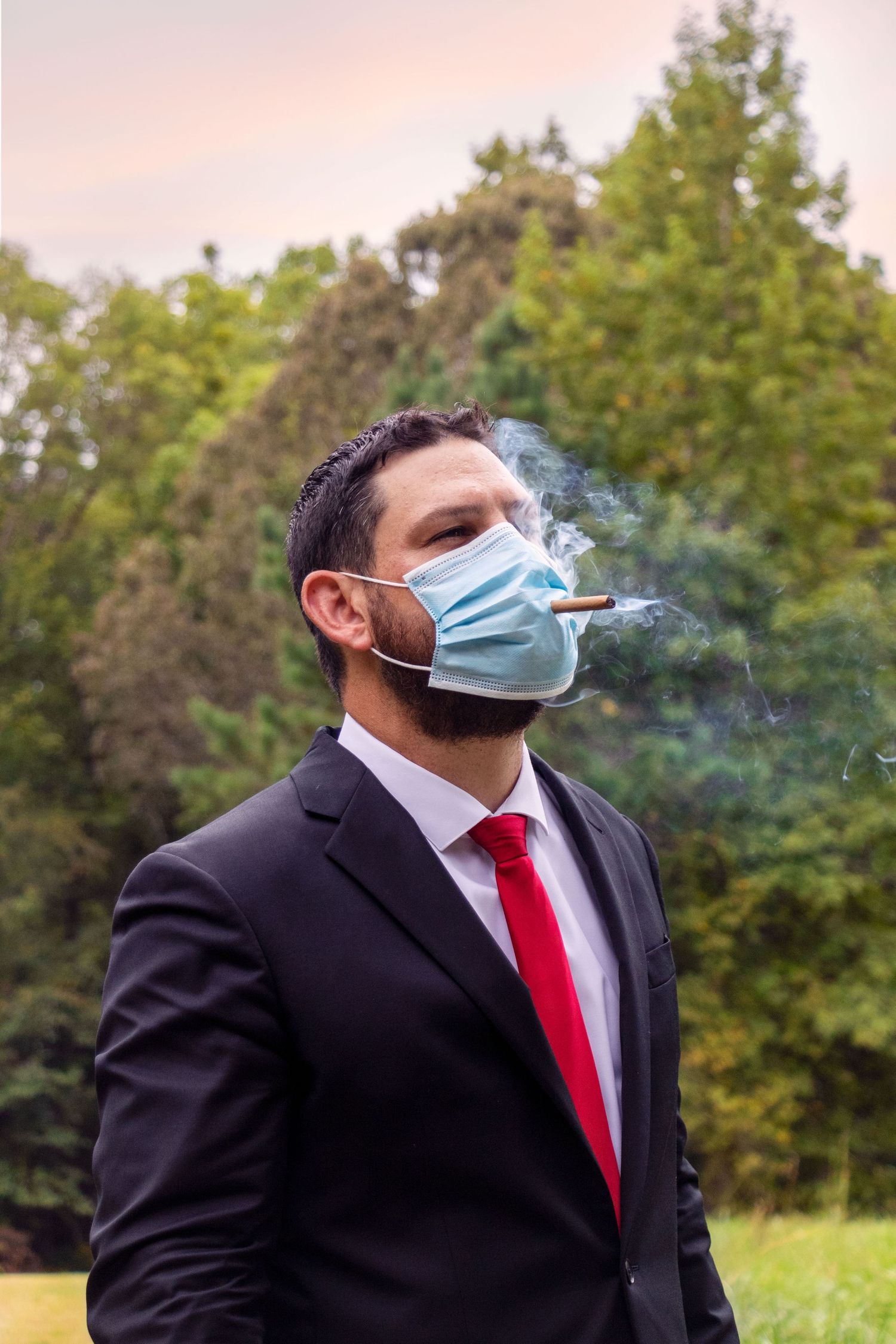 A suited man in a face mask smokes a cigar outdoors with trees in the background.