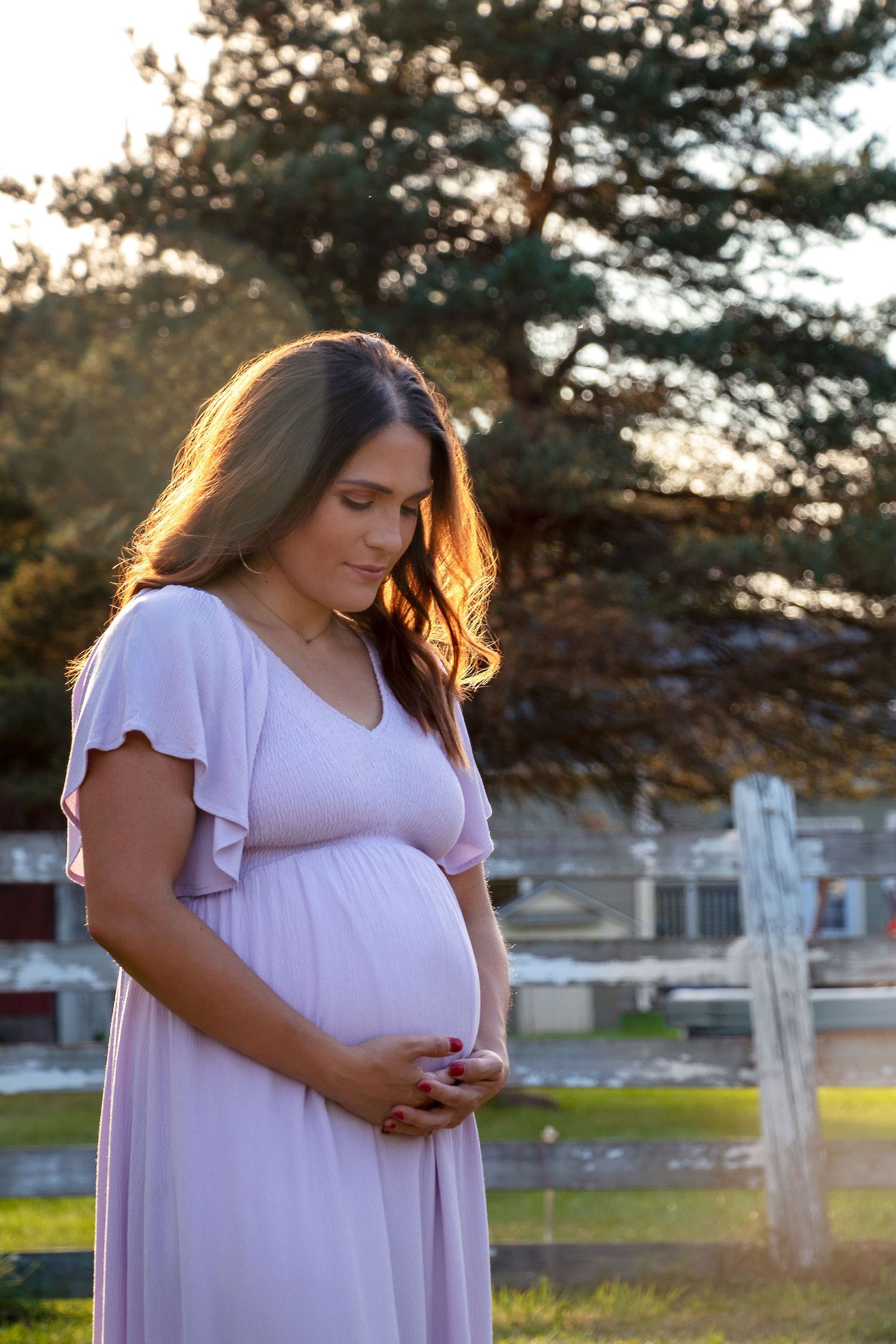 A glowing sunset portrait of an expectant mother in a flowing pink dress standing in an outdoor setting.