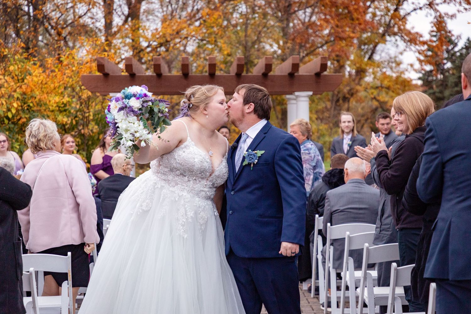 Outdoor autumn wedding ceremony with guests seated in white chairs surrounded by fall foliage.