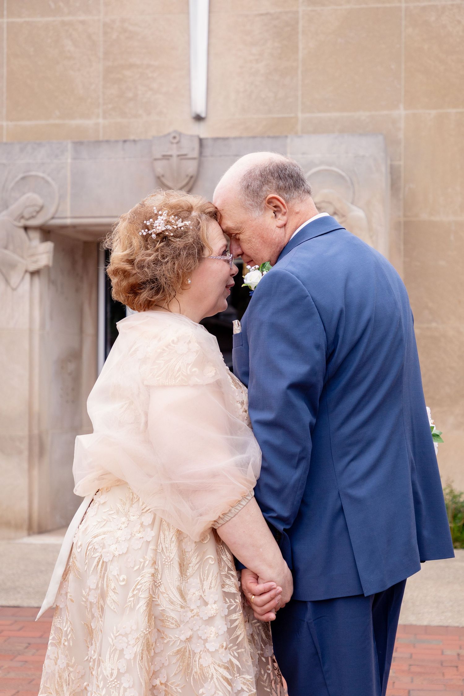 A couple shares a tender moment in front of a stone building wall wearing formal attire.