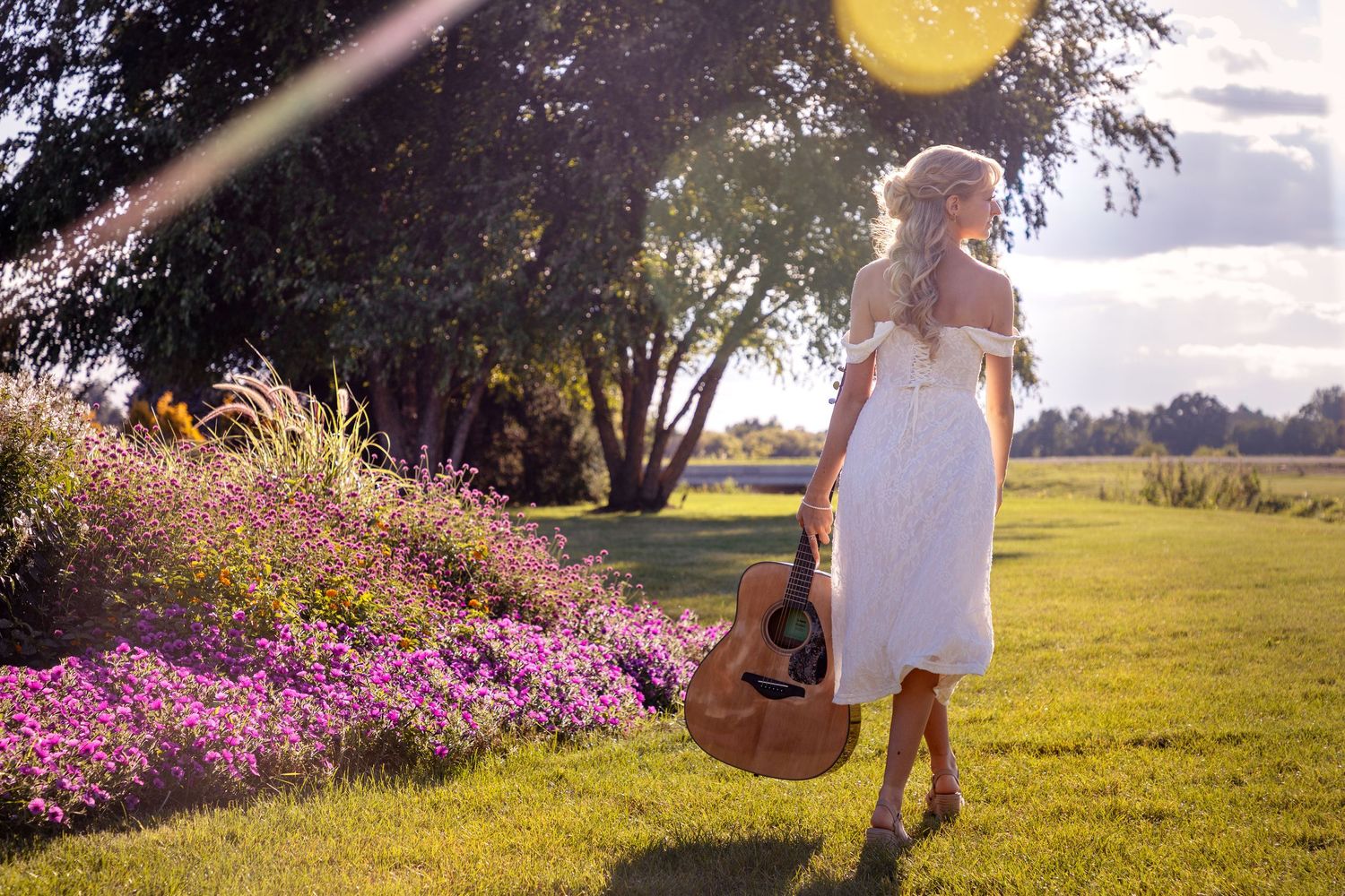 Person in white dress walking through sunlit field with wildflowers.