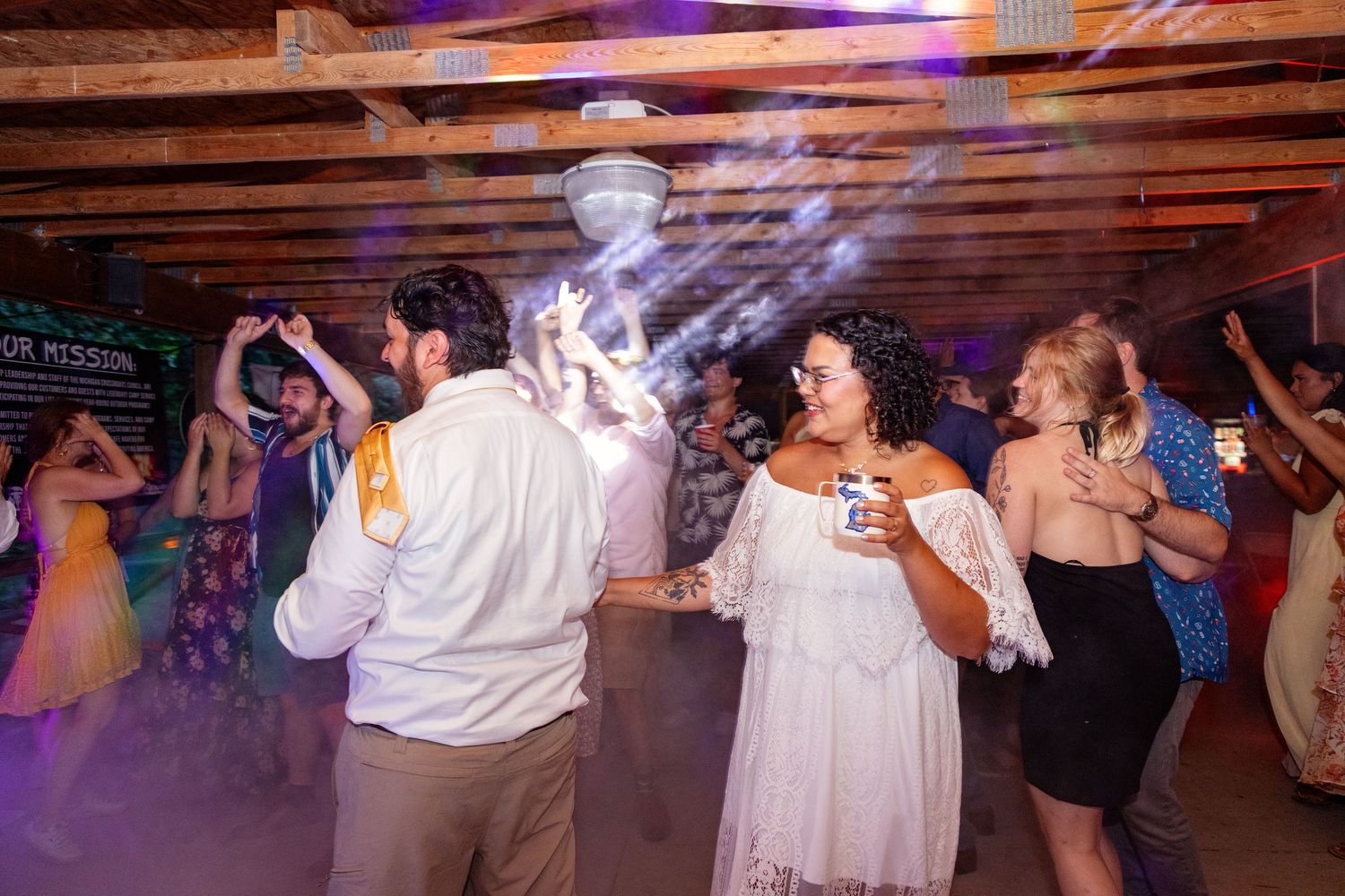 Wedding reception dance party with colorful lights and smoke effects in a rustic venue.