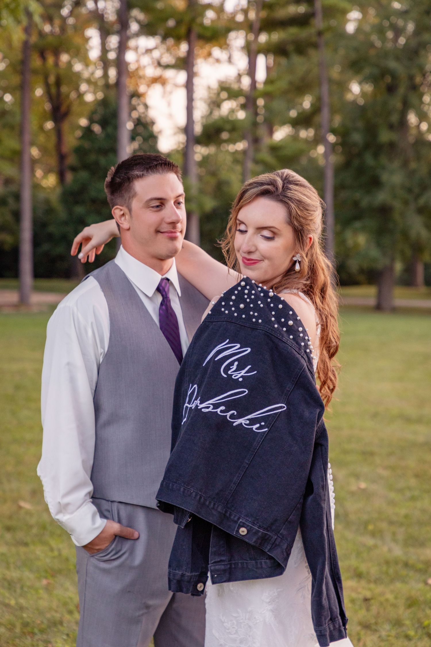 Couple poses in casual wedding attire with custom denim jacket in park setting.