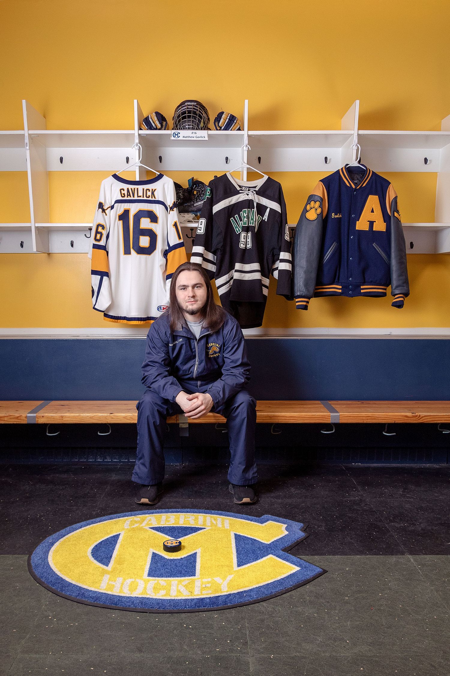 Student sits in hockey locker room with jerseys and equipment displayed on wall.